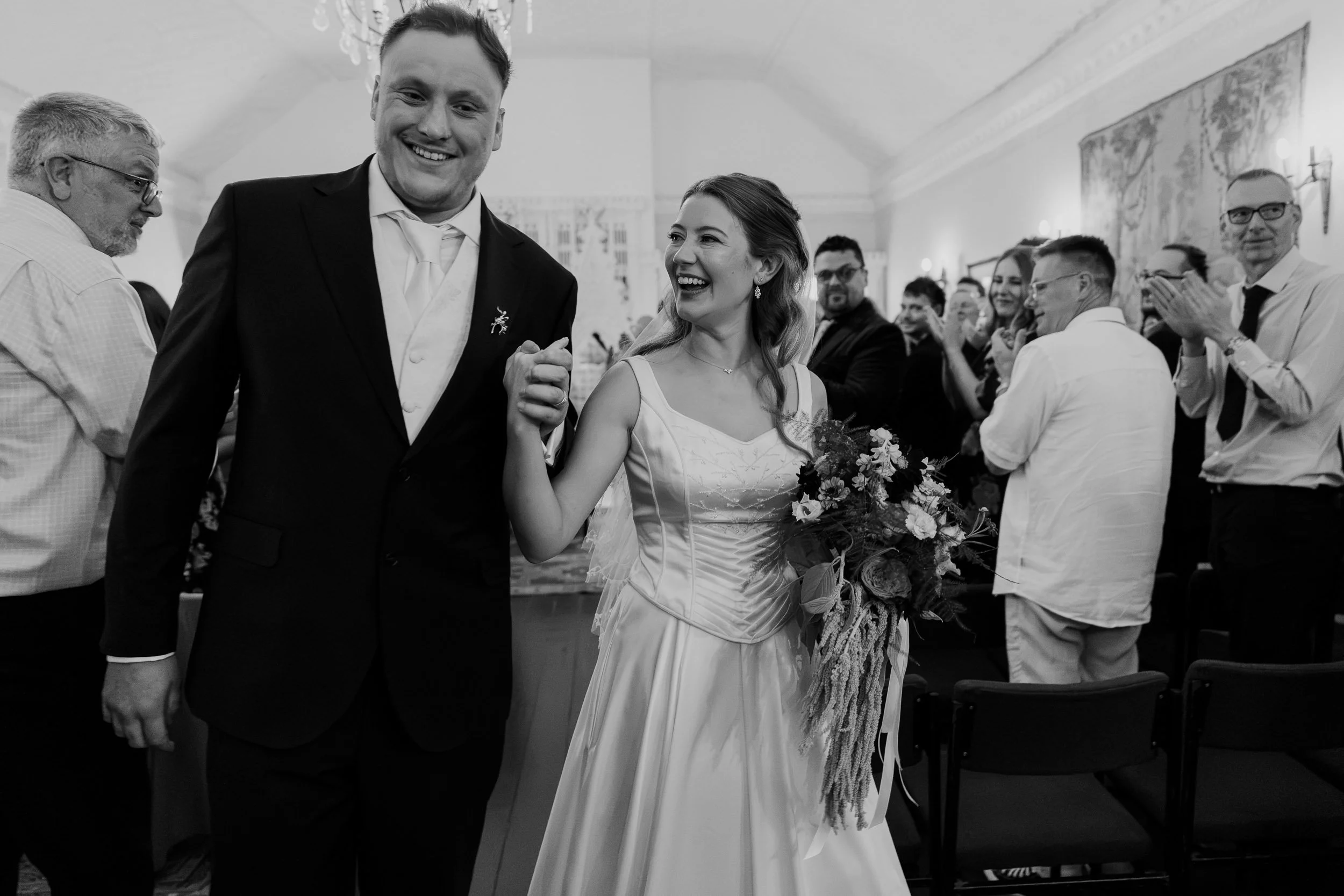 A joyful bride and groom holding hands and smiling at a wedding reception, surrounded by seated guests clapping and enjoying the celebration.