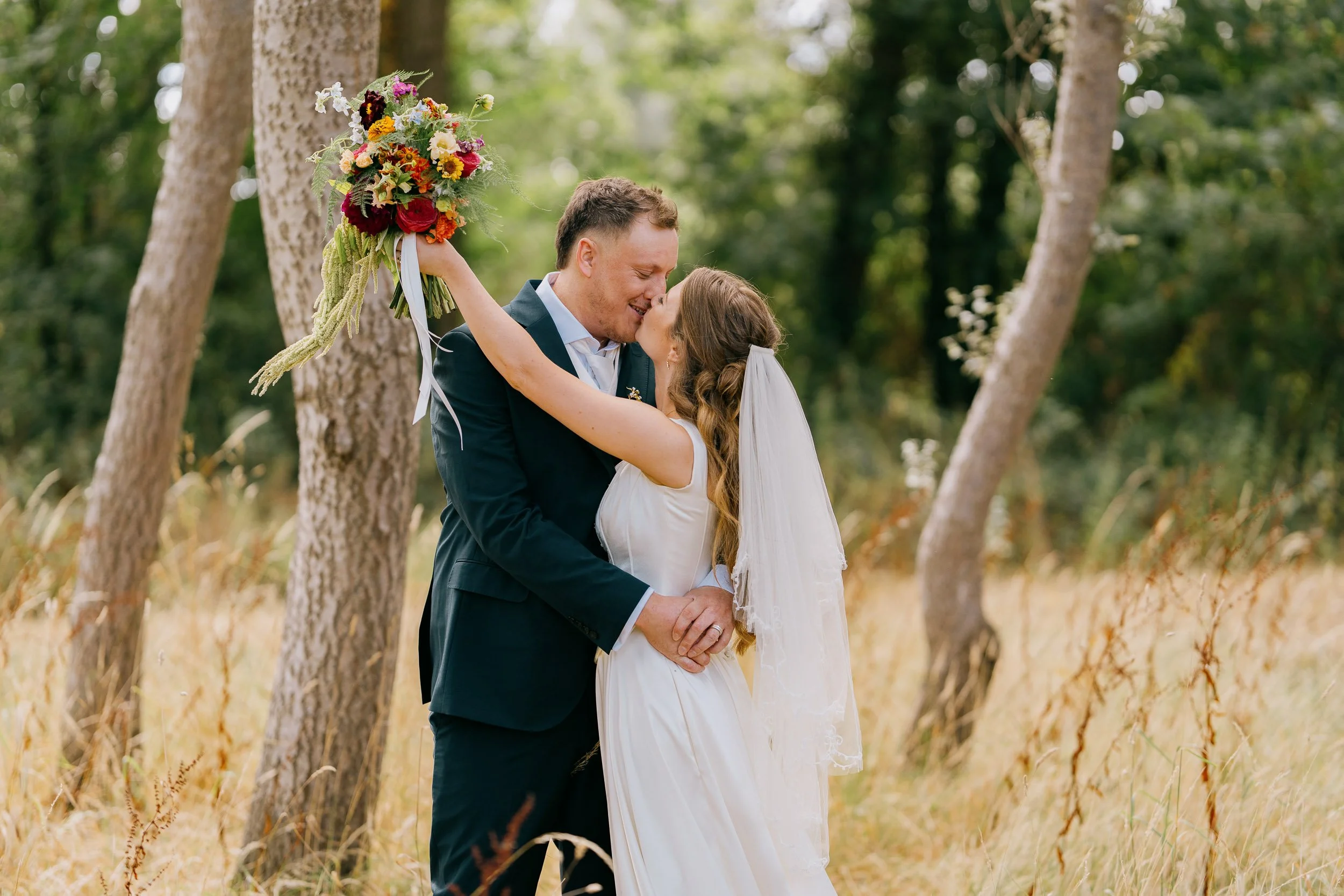 A bride and groom sharing a kiss outdoors during their wedding ceremony, with the bride holding a colorful bouquet and surrounded by trees.