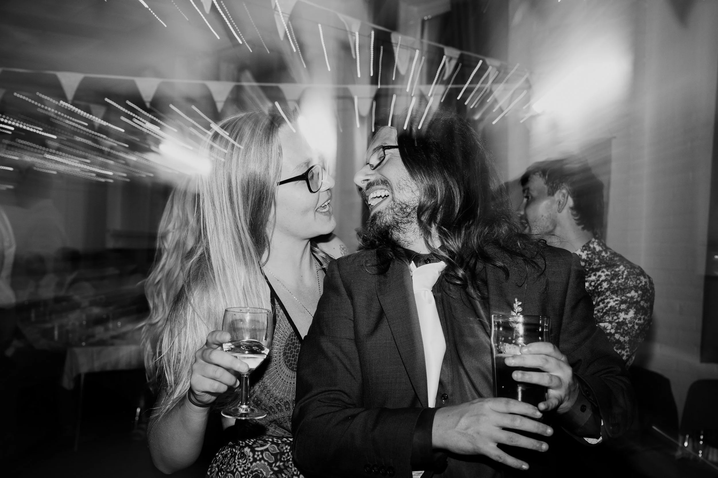 Two women and a man are smiling and holding drinks at a celebration or party. They are close together, with the women leaning in towards each other and the man. The background has blurred lights and other people, indicating a lively social event.