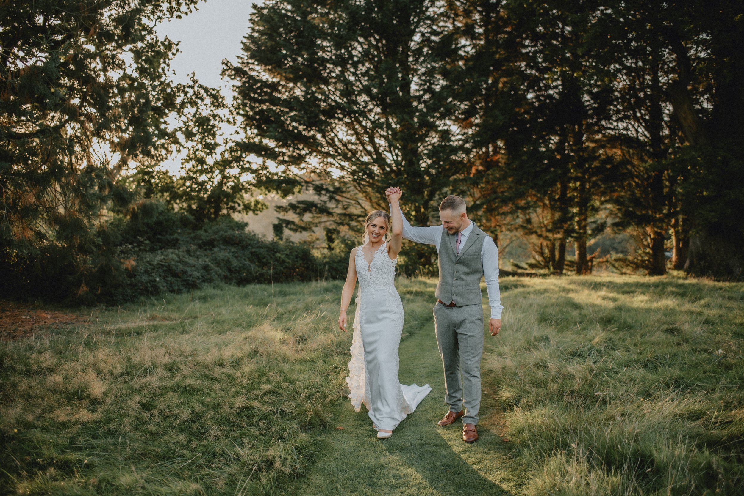A bride and groom are walking hand in hand on a grassy path through a park or forest during sunset, with the bride wearing a white wedding gown and the groom in a light gray suit.
