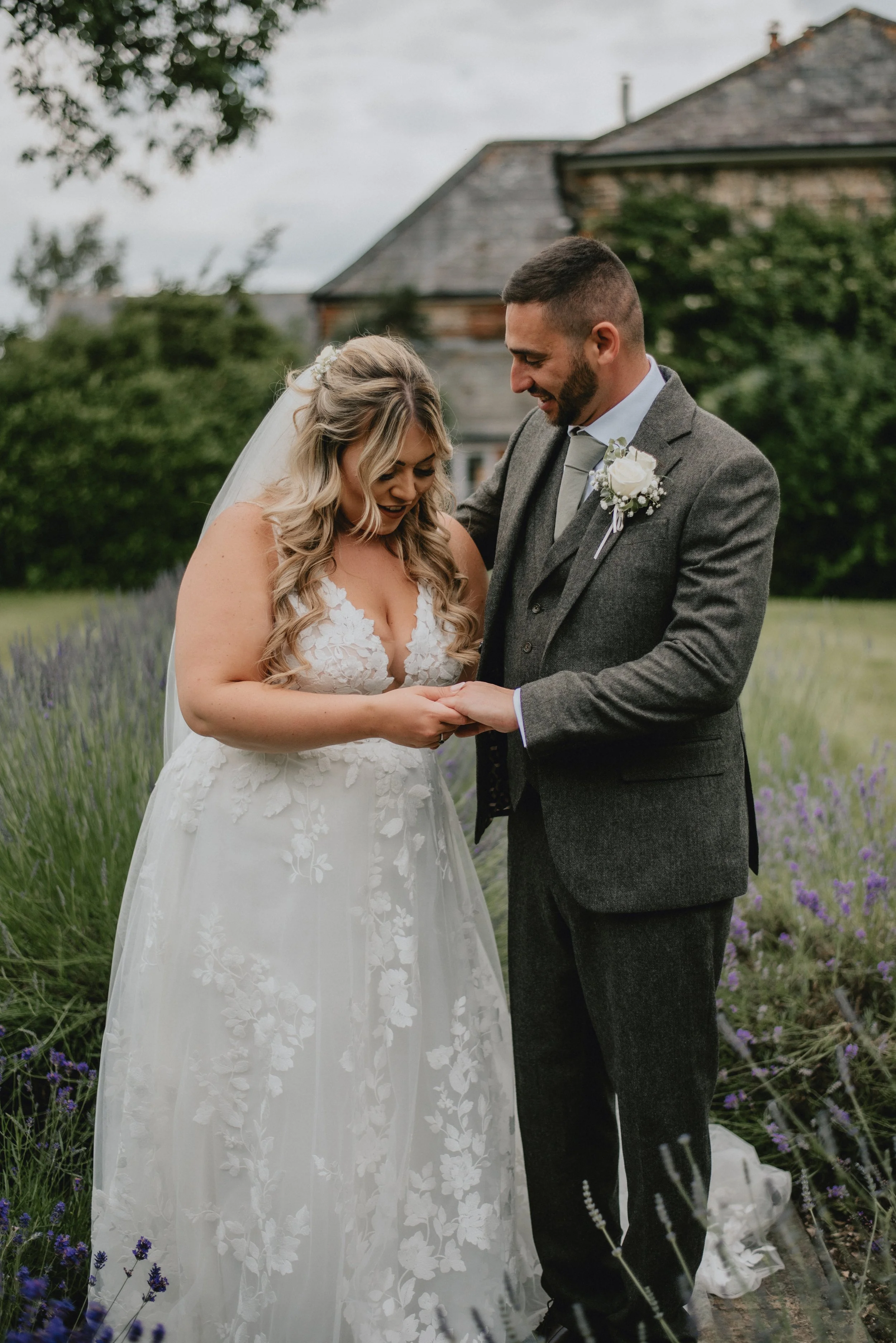 Bride and groom exchange vows in a lavender field, with the bride in a white lace gown and the groom in a gray suit, outdoors with a house and trees in the background.