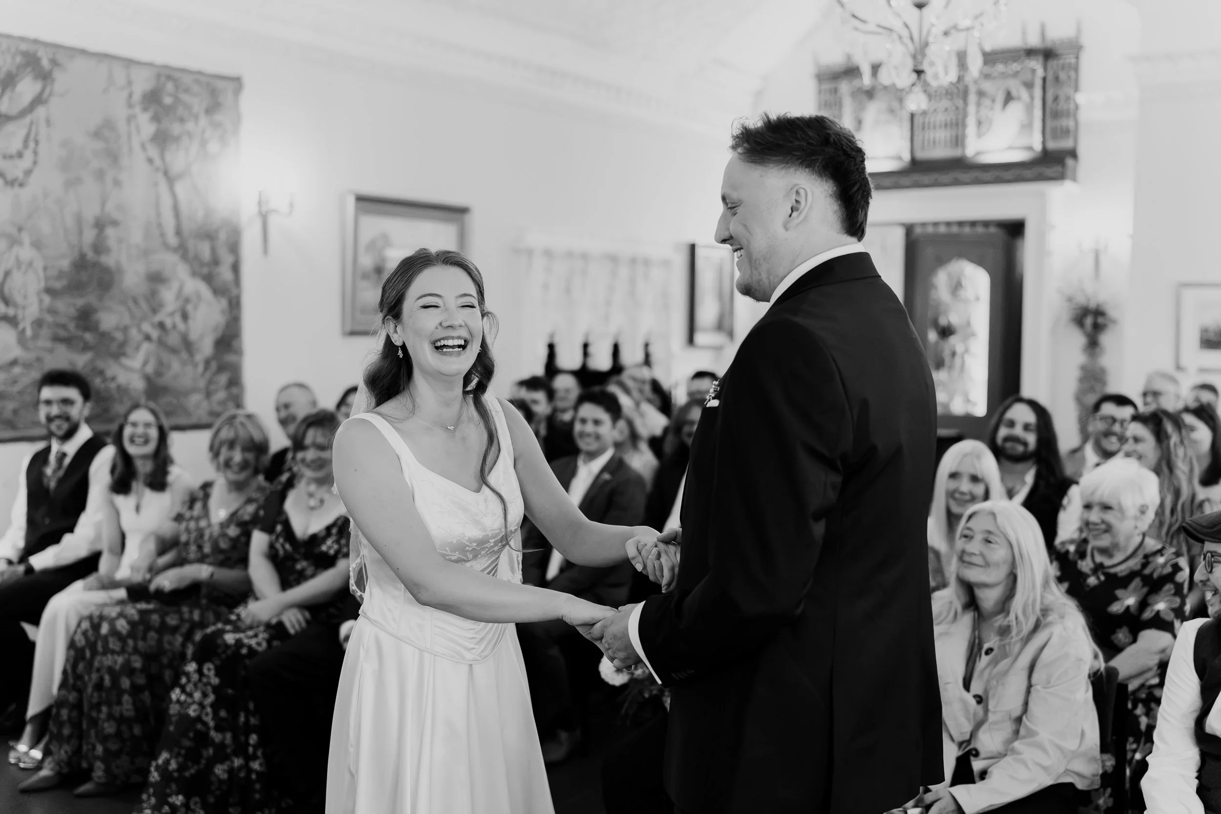 A black and white photo of a bride and groom holding hands and smiling during their wedding ceremony inside a decorated hall, with guests seated in the background.