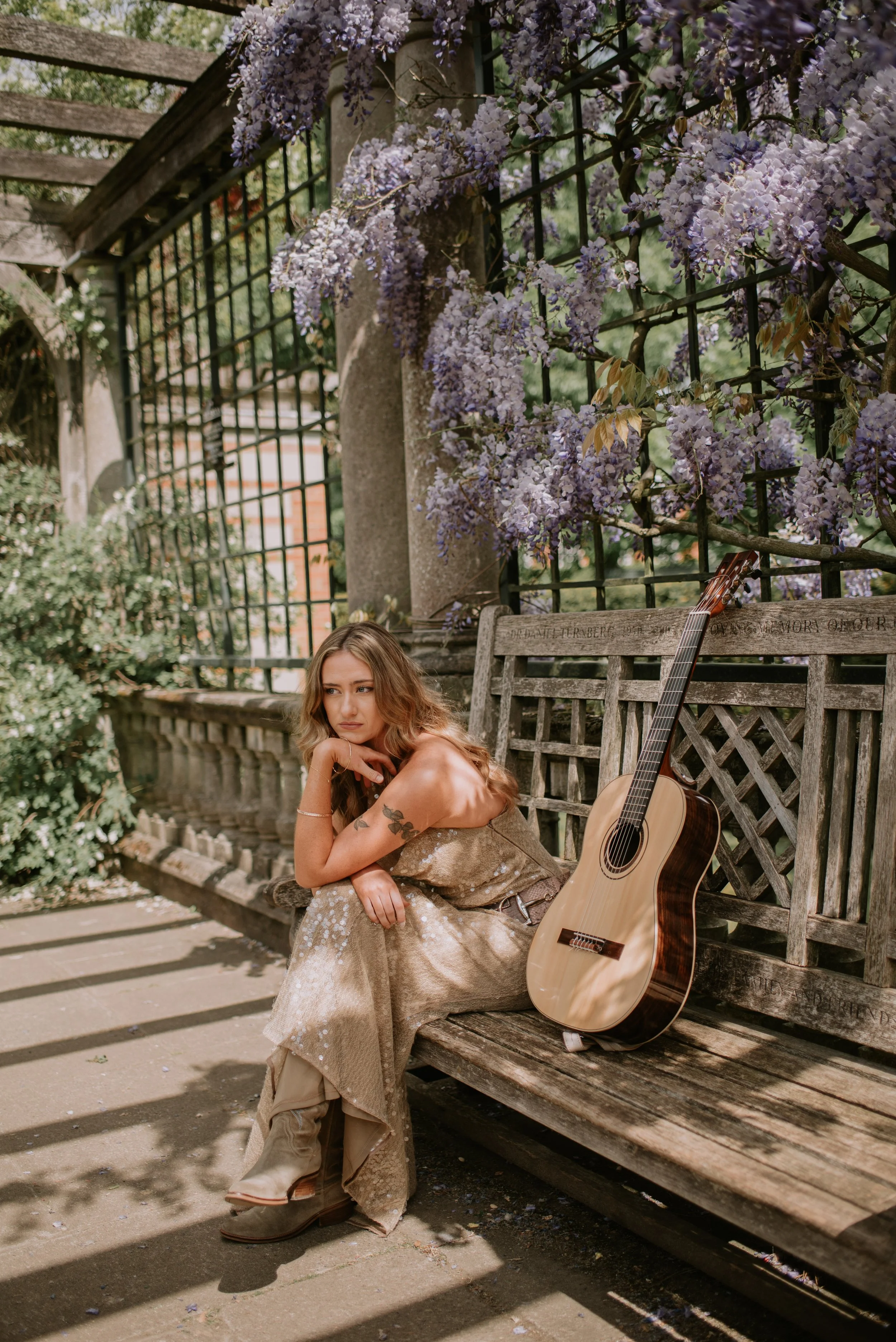Young woman sitting on a wooden bench with a guitar beside her, surrounded by purple wisteria flowers on a sunny day in a garden.