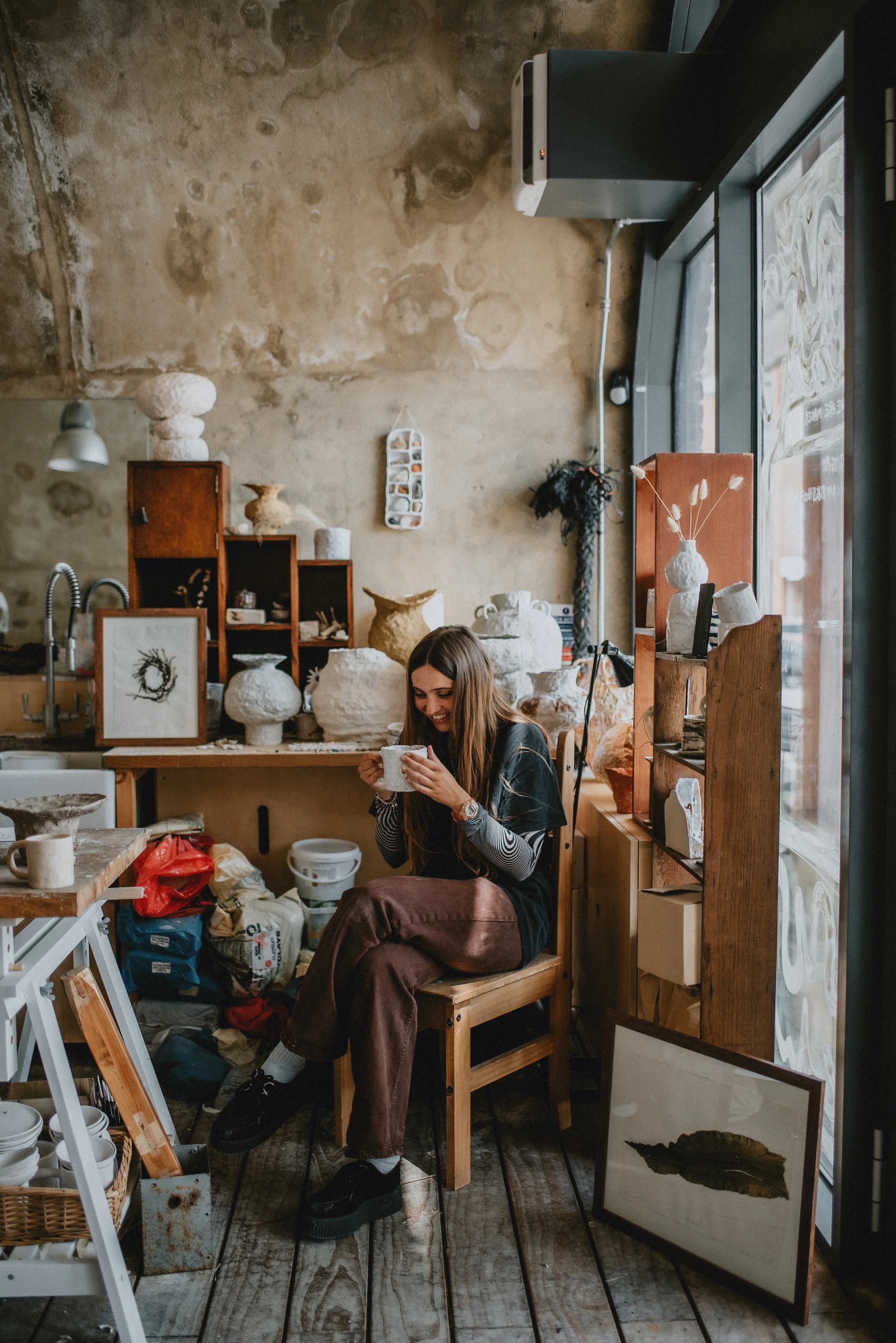 A woman seated on a wooden chair inside an art studio or ceramic workspace, holding a mug and smiling, with pottery and art supplies around her and natural light coming through large windows.