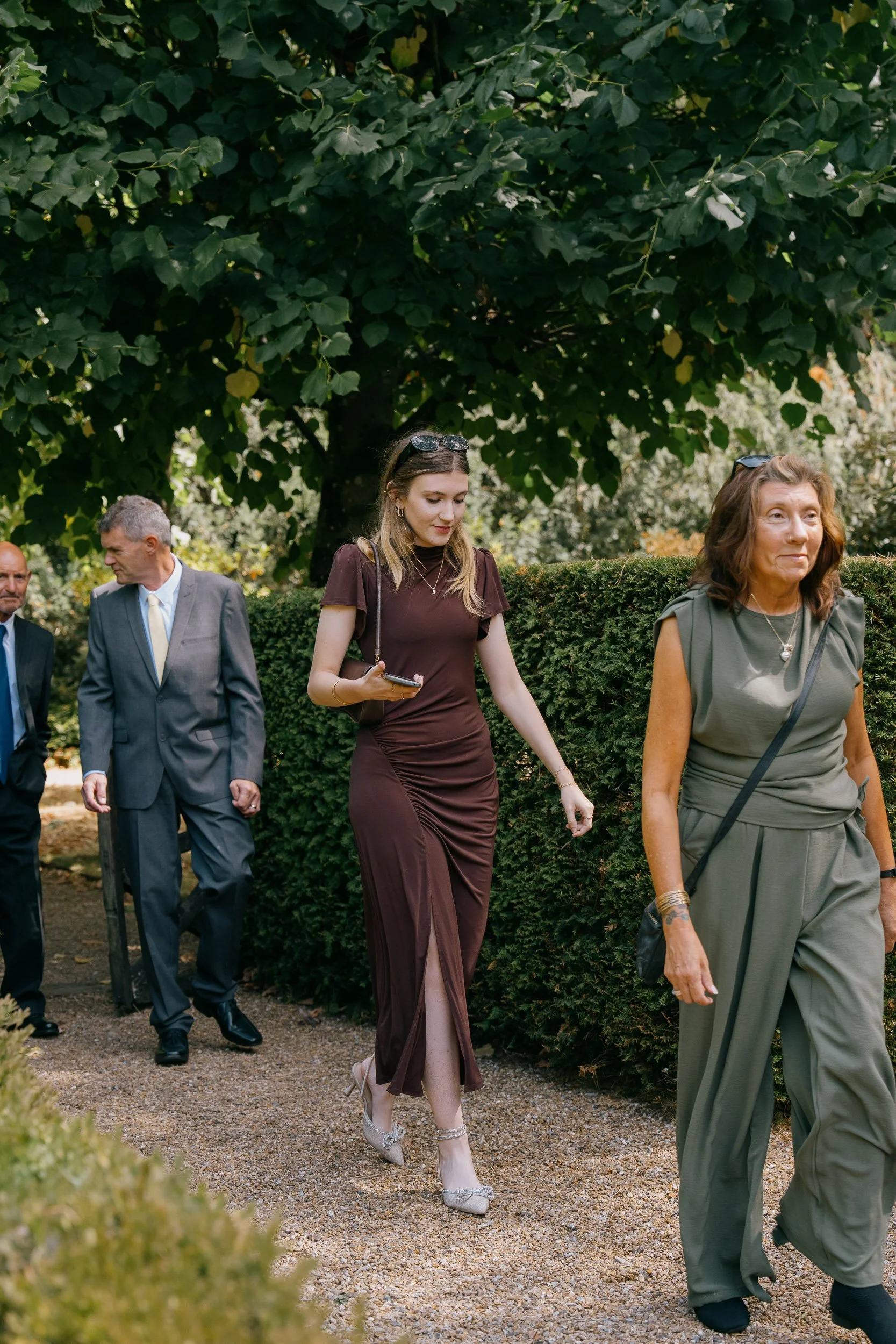 A group of people walking on a gravel path, with a large leafy tree and green hedge in the background. The group consists of men and women dressed in formal and semi-formal attire, engaged in conversation.