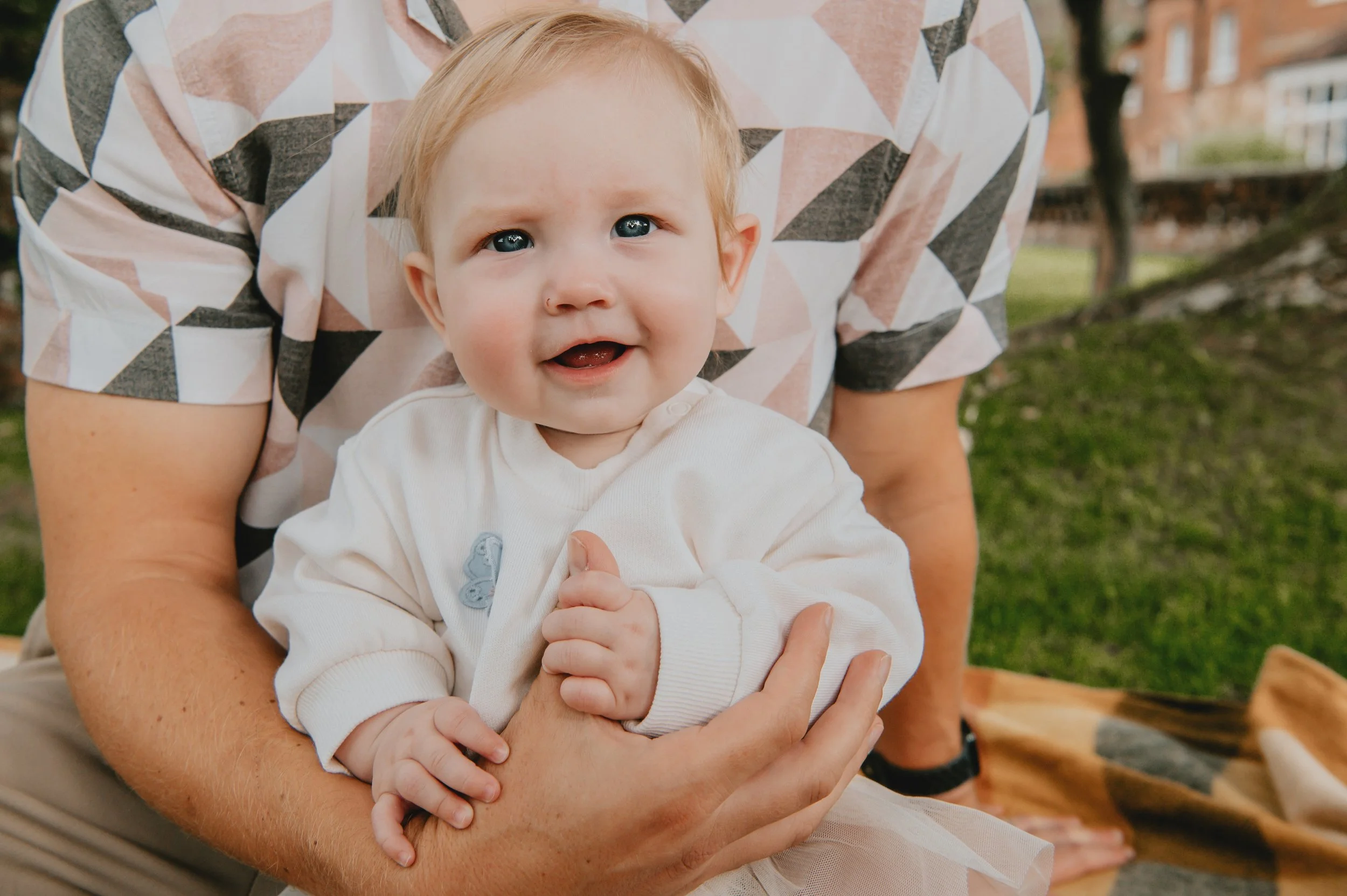 A baby with blue eyes and light hair, wearing a cream-colored sweater, is being held by an adult. The person, wearing a multicolored geometric pattern shirt, is crouched down outdoors on a blanket in a grassy area with trees and buildings in the background.