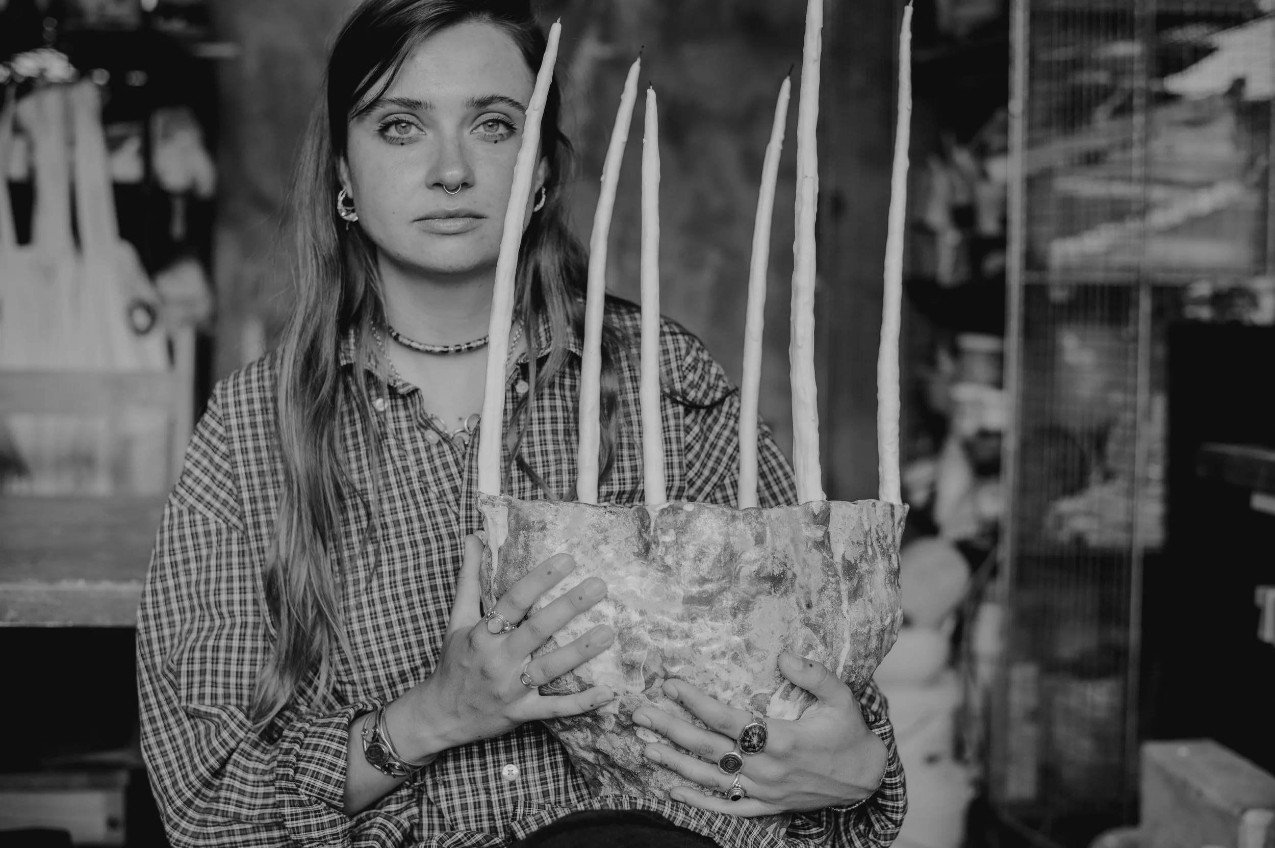 A woman with long hair and tattoos on her fingers holding a cake with lit candles in a store or workshop setting.