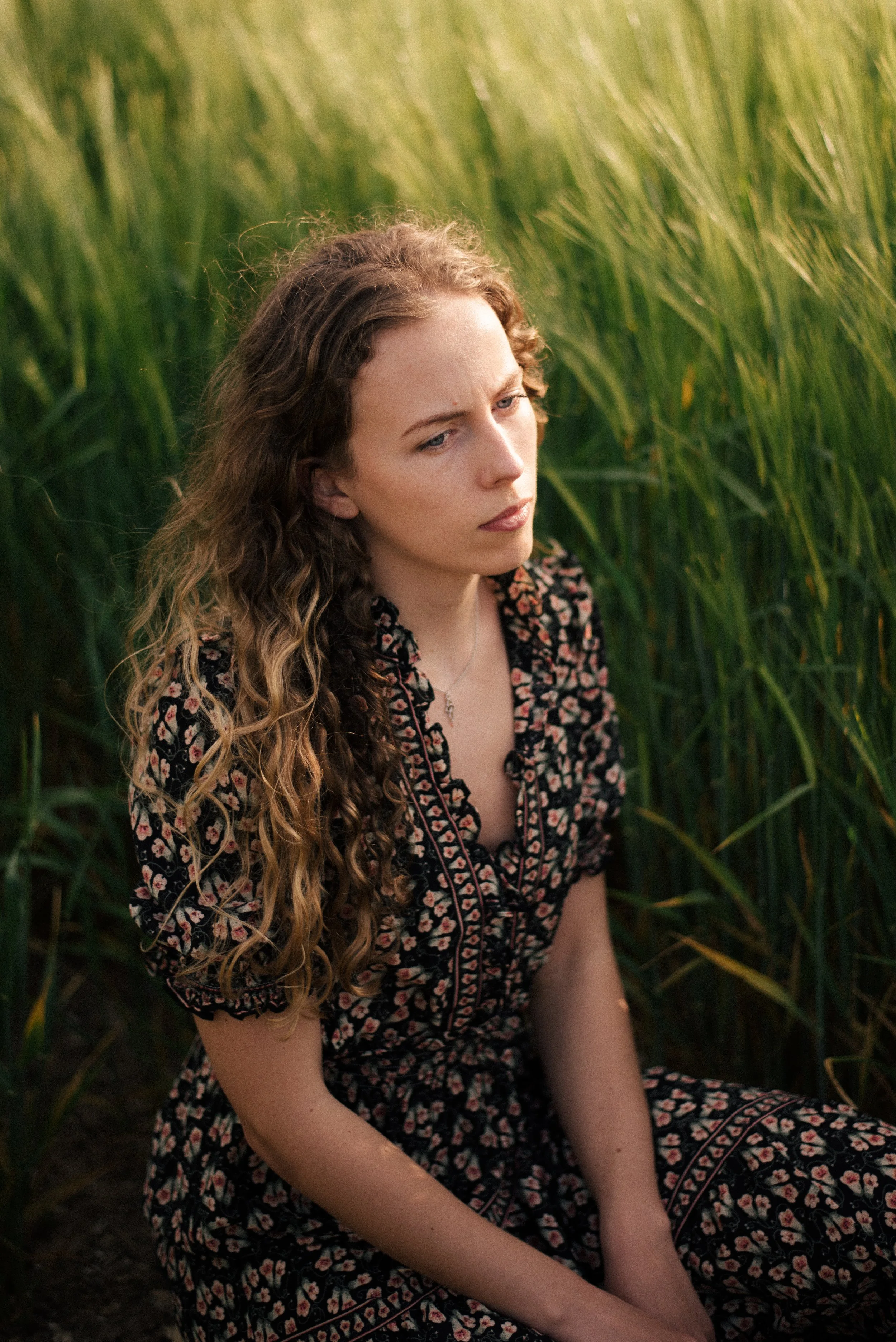 A young woman with curly blonde hair sitting in a field of tall green grass, looking contemplative.