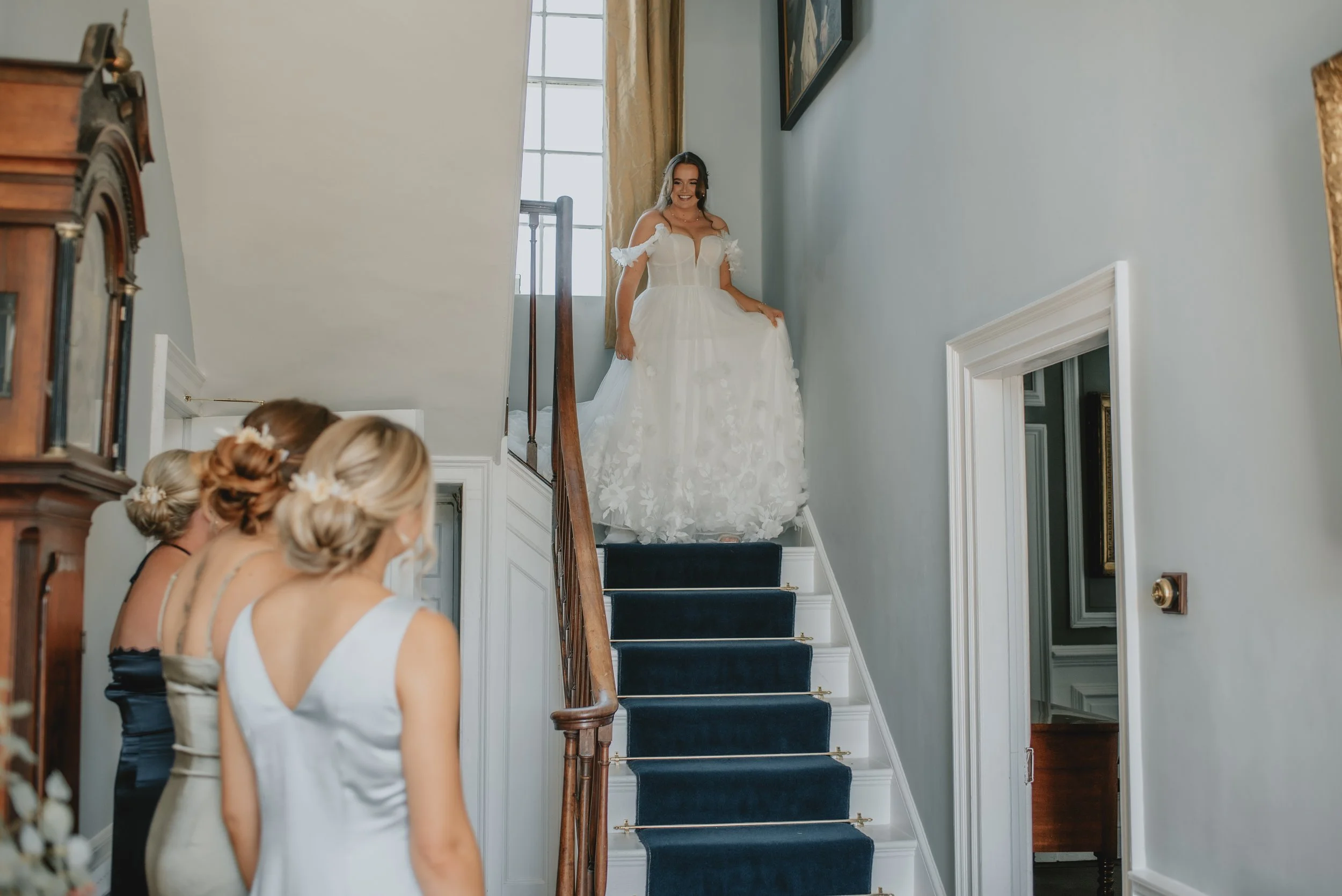 Bride wearing a white wedding dress standing on staircase with dark blue carpet, smiling at three women in formal dresses at the bottom of the stairs.