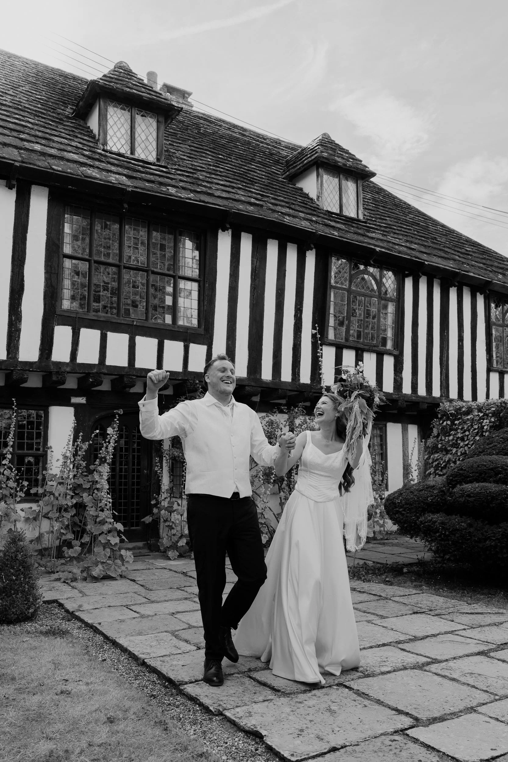 A smiling man and woman in wedding attire holding hands and celebrating outdoors in front of a half-timbered cottage, with a garden and pathway.