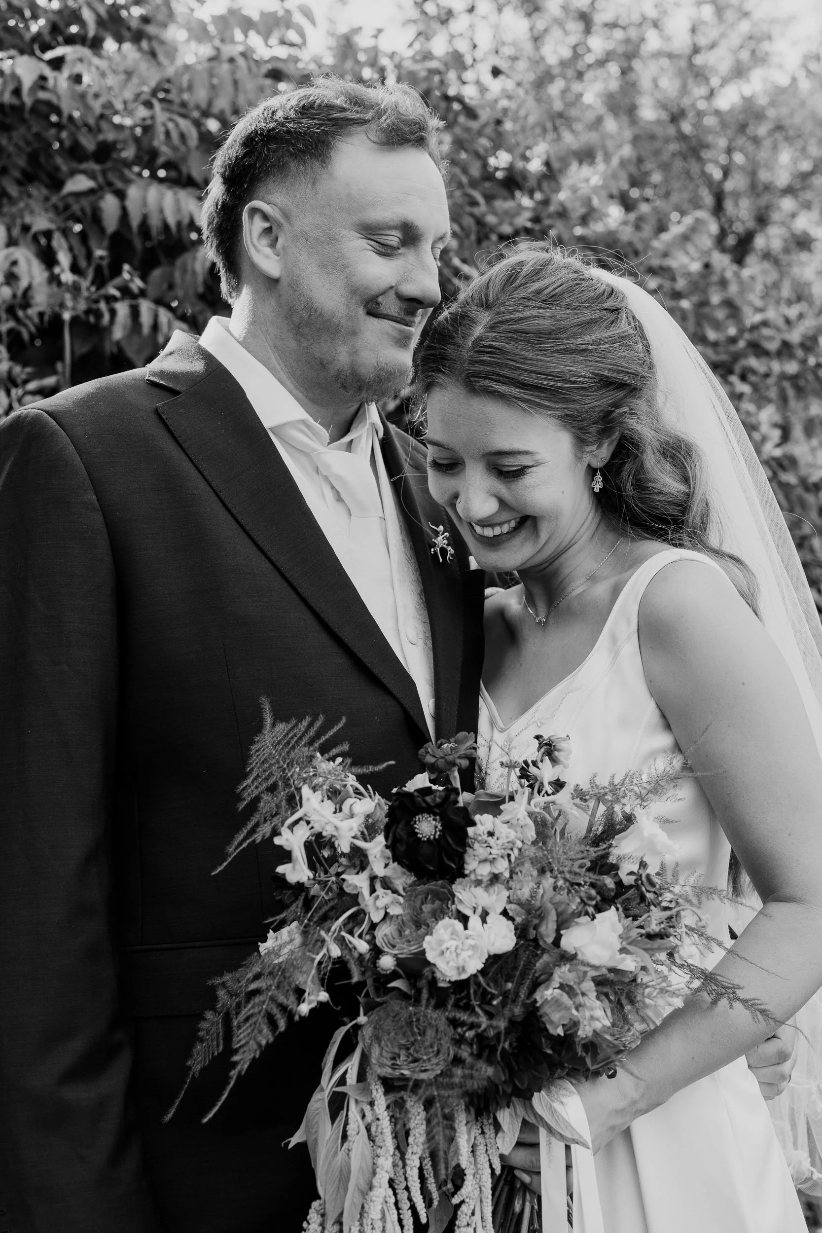 A black and white photo of a happy couple, likely at their wedding, standing close and smiling with eyes closed. The man wears a dark suit with a light shirt and a bow tie, and the woman wears a white dress with a veil and earrings, holding a large b