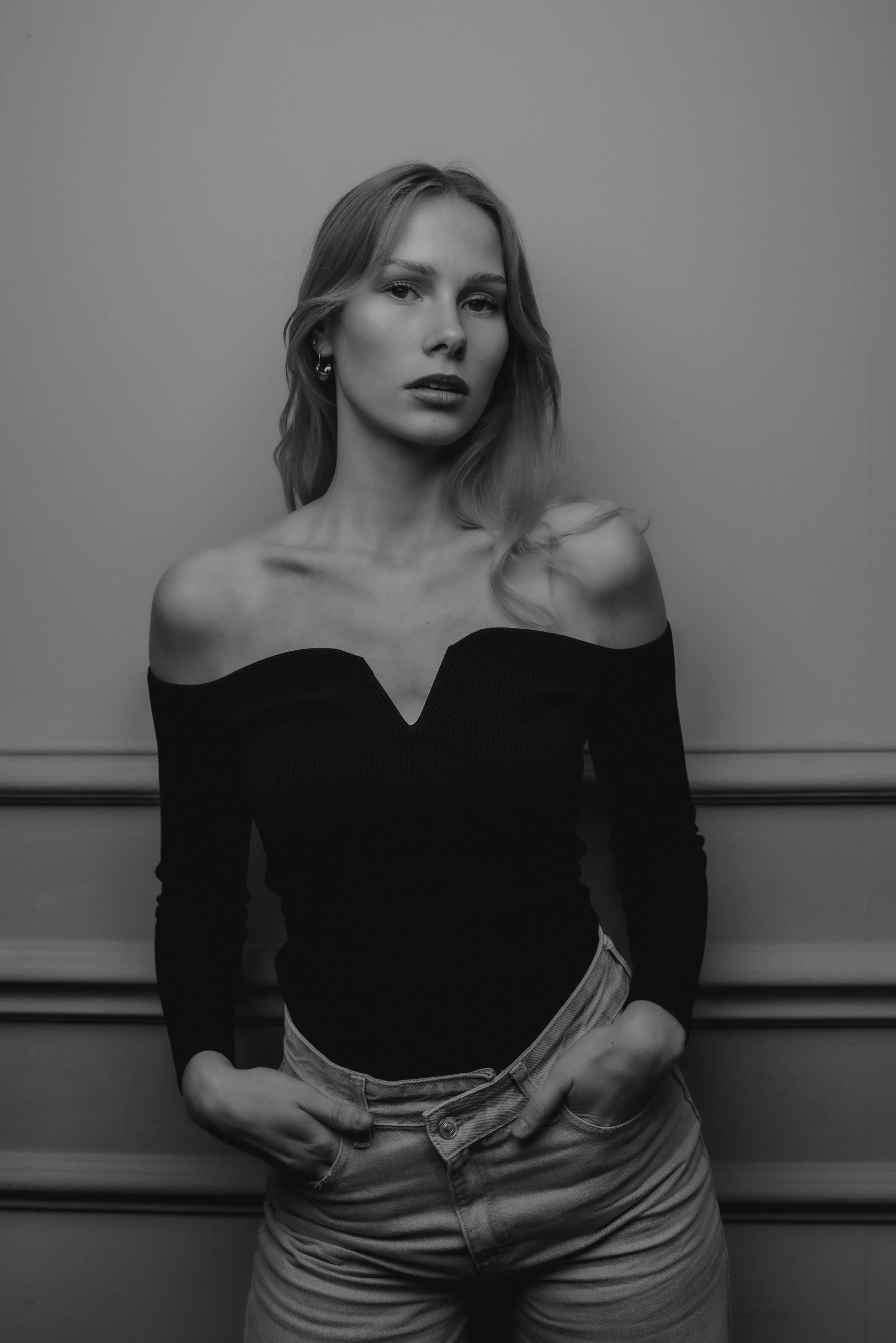 A black and white photo of a young woman with blonde hair, wearing a off-the-shoulder black top and light-colored jeans, posing with her hands in her pockets against a simple background.