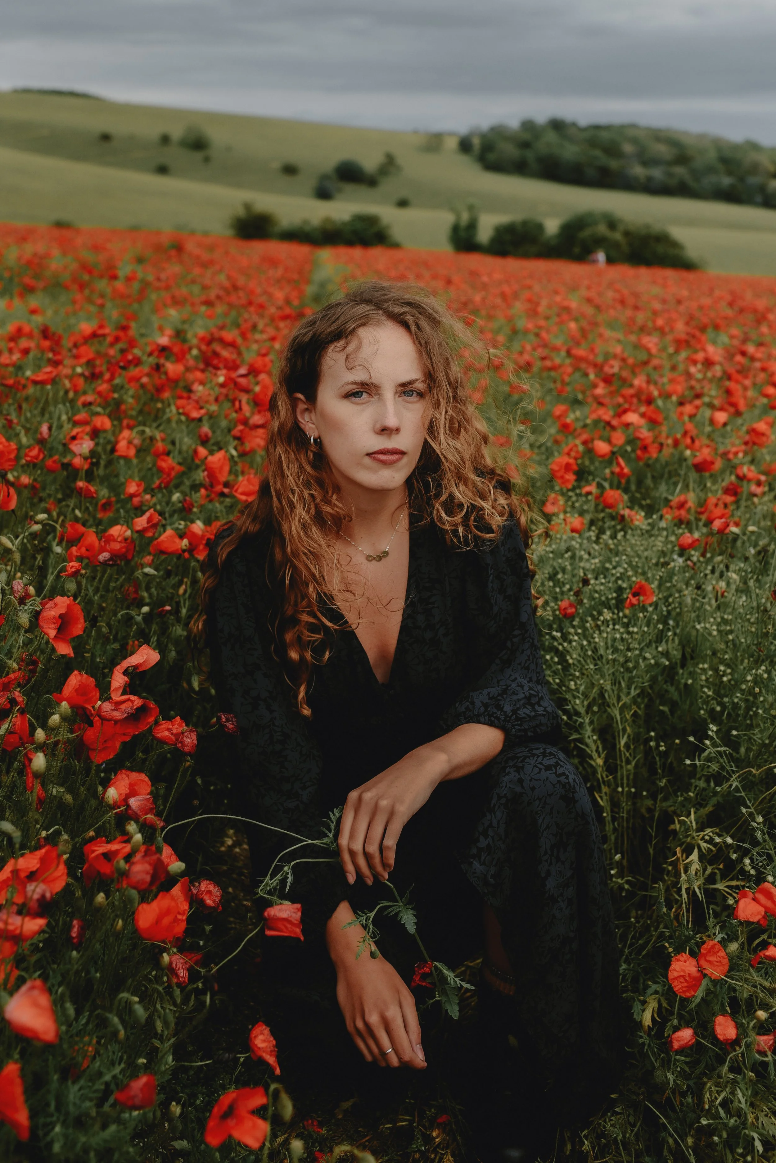 A woman with curly hair wearing a black dress sitting among red poppy flowers in a field with green rolling hills and a cloudy sky in the background.