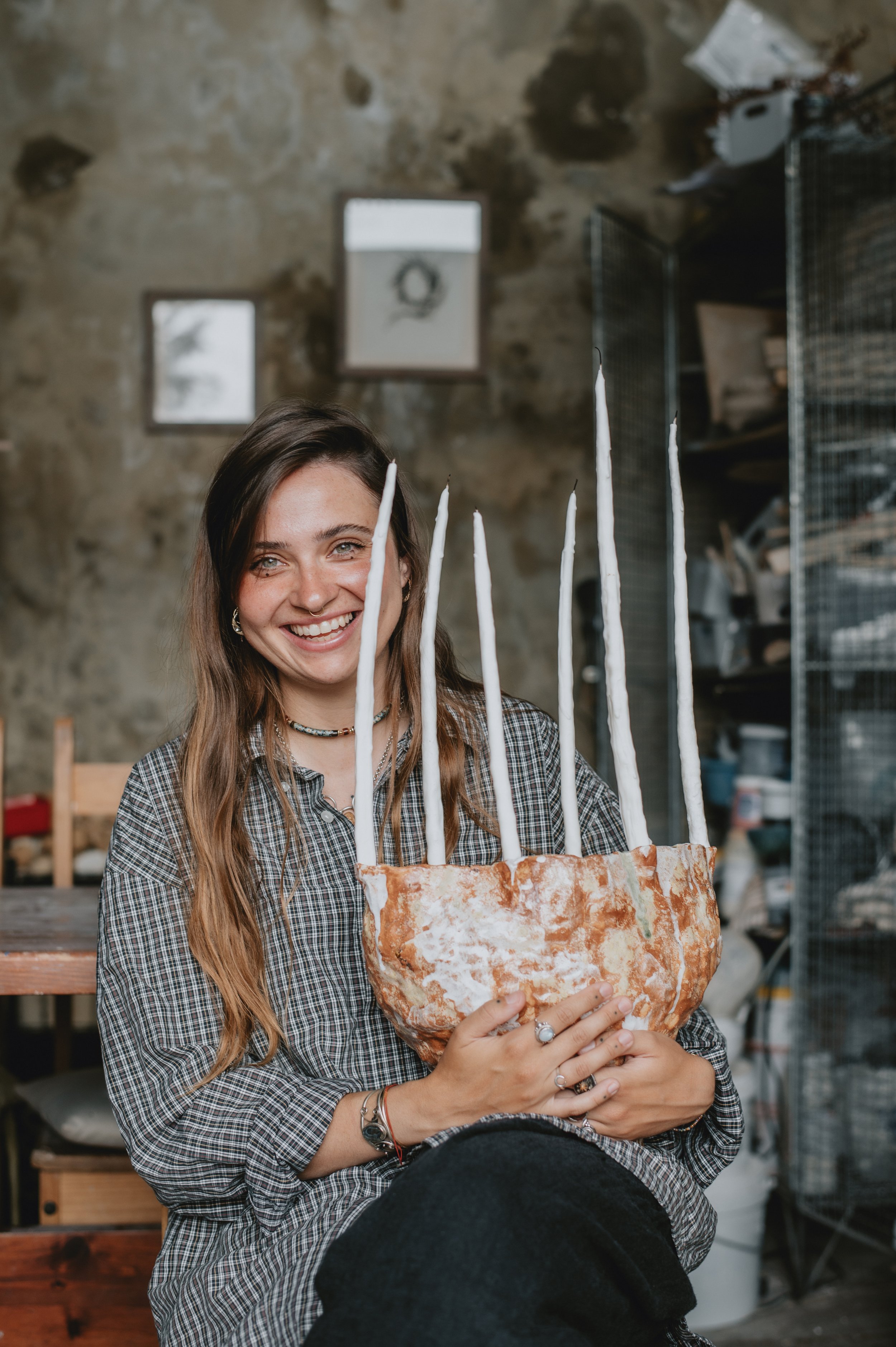 A young woman with long brown hair and a checkered shirt is sitting and smiling while holding a large cake with seven white candles. The background features a rustic, industrial-style interior with shelves and framed pictures on a stone wall.