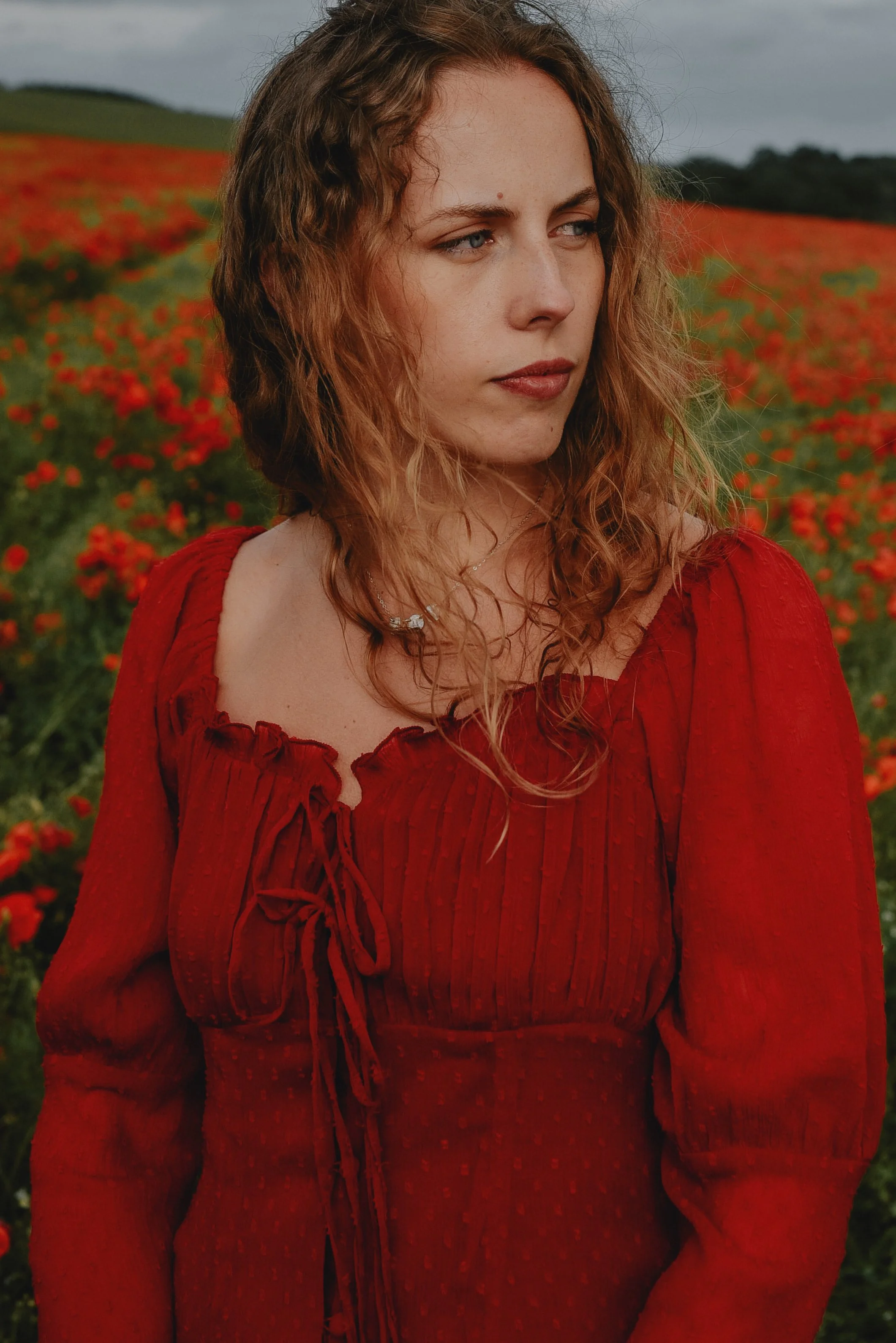 A woman with wavy, reddish-brown hair wearing a red dress stands in a field of red flowers, looking to her left with a contemplative expression.