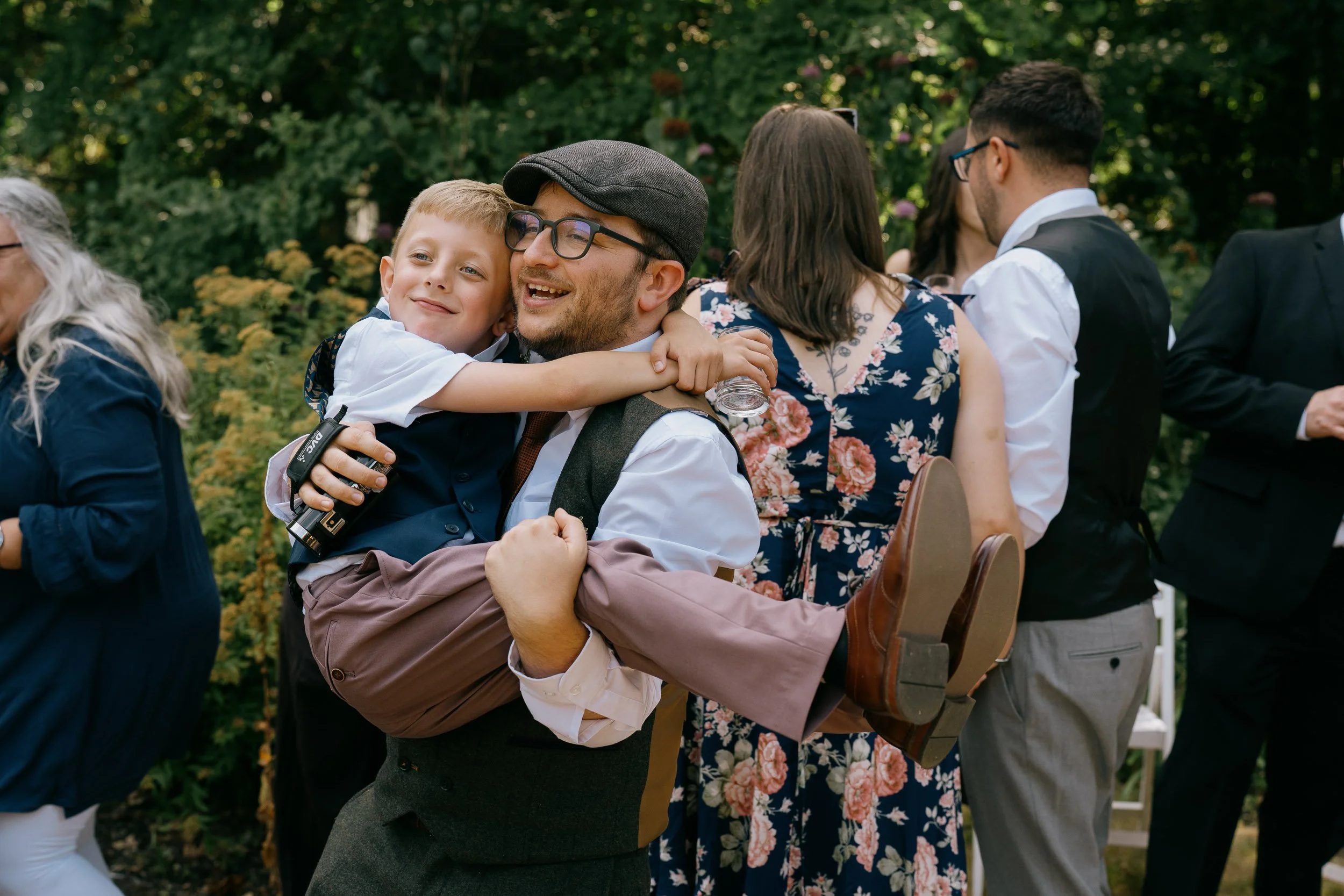 A man with glasses, a cap, and a vest holds a young boy in a white shirt as they embrace and smile at an outdoor gathering in a garden setting.
