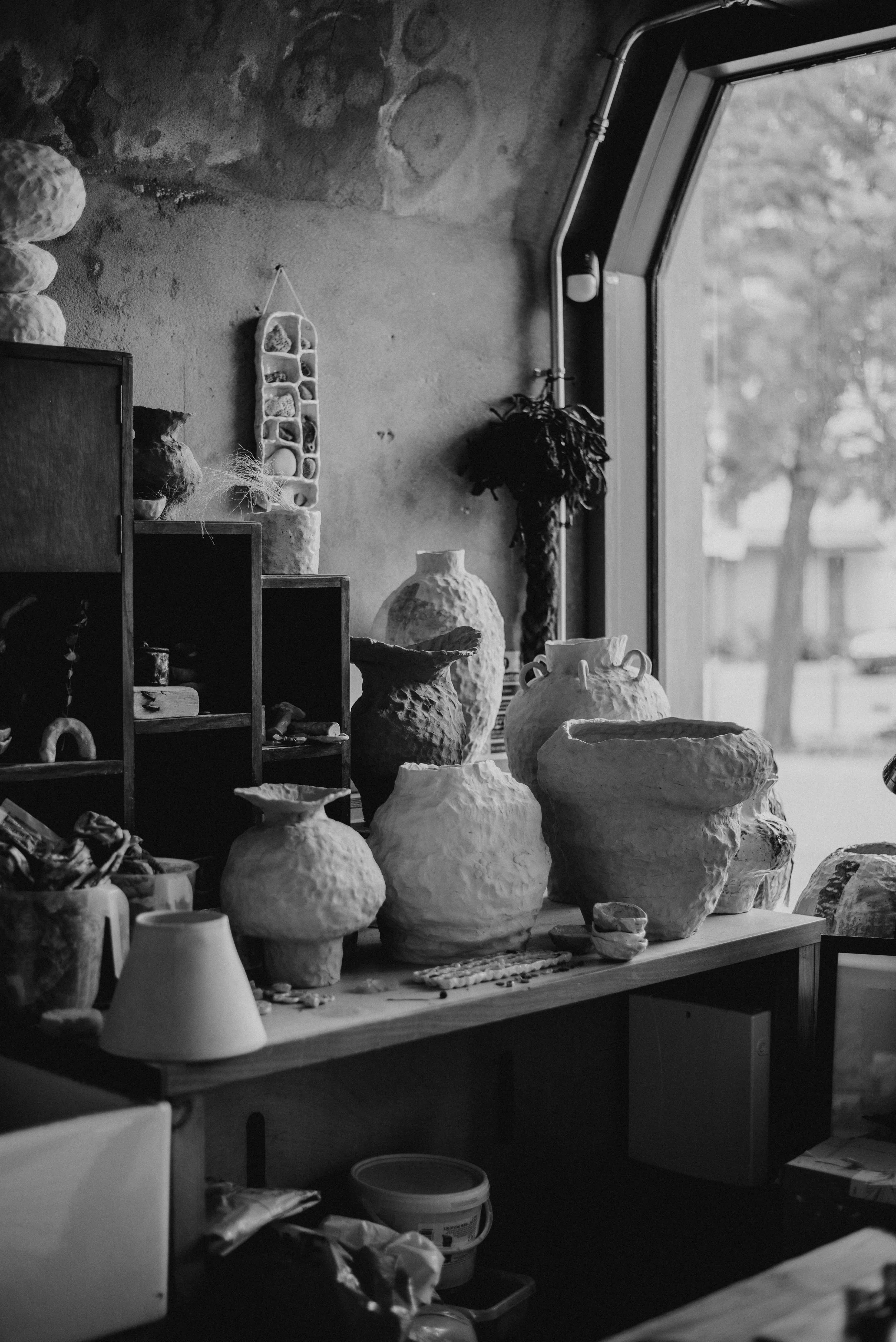A collection of unglazed ceramic vases and pots on a wooden table inside a pottery studio, with a large window in the background showing trees outside.