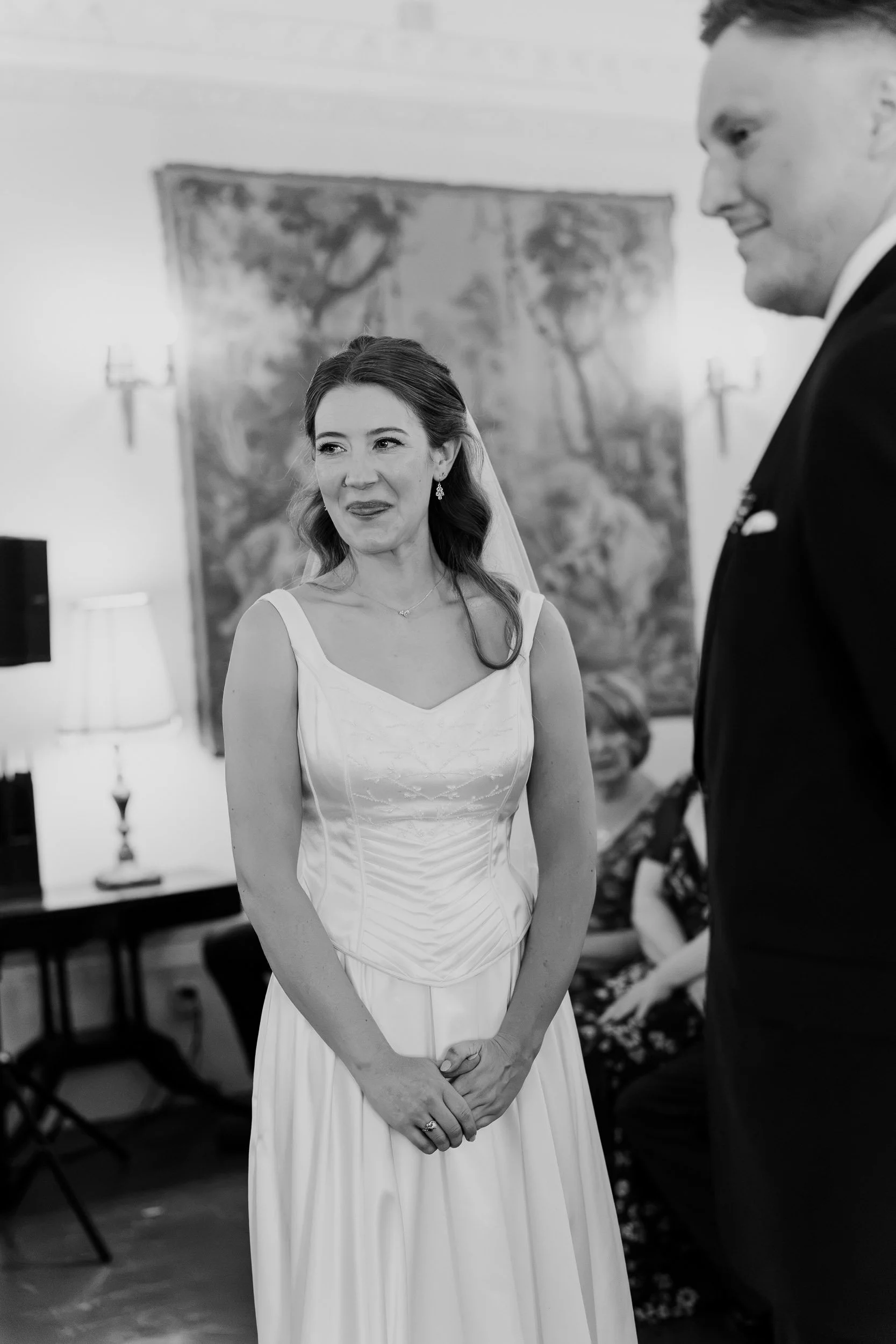 A bride in a white wedding dress with her hands clasped in front of her, smiling softly, standing indoors with a painting and a lamp in the background. A man in a suit is partially visible on the right side of the image.
