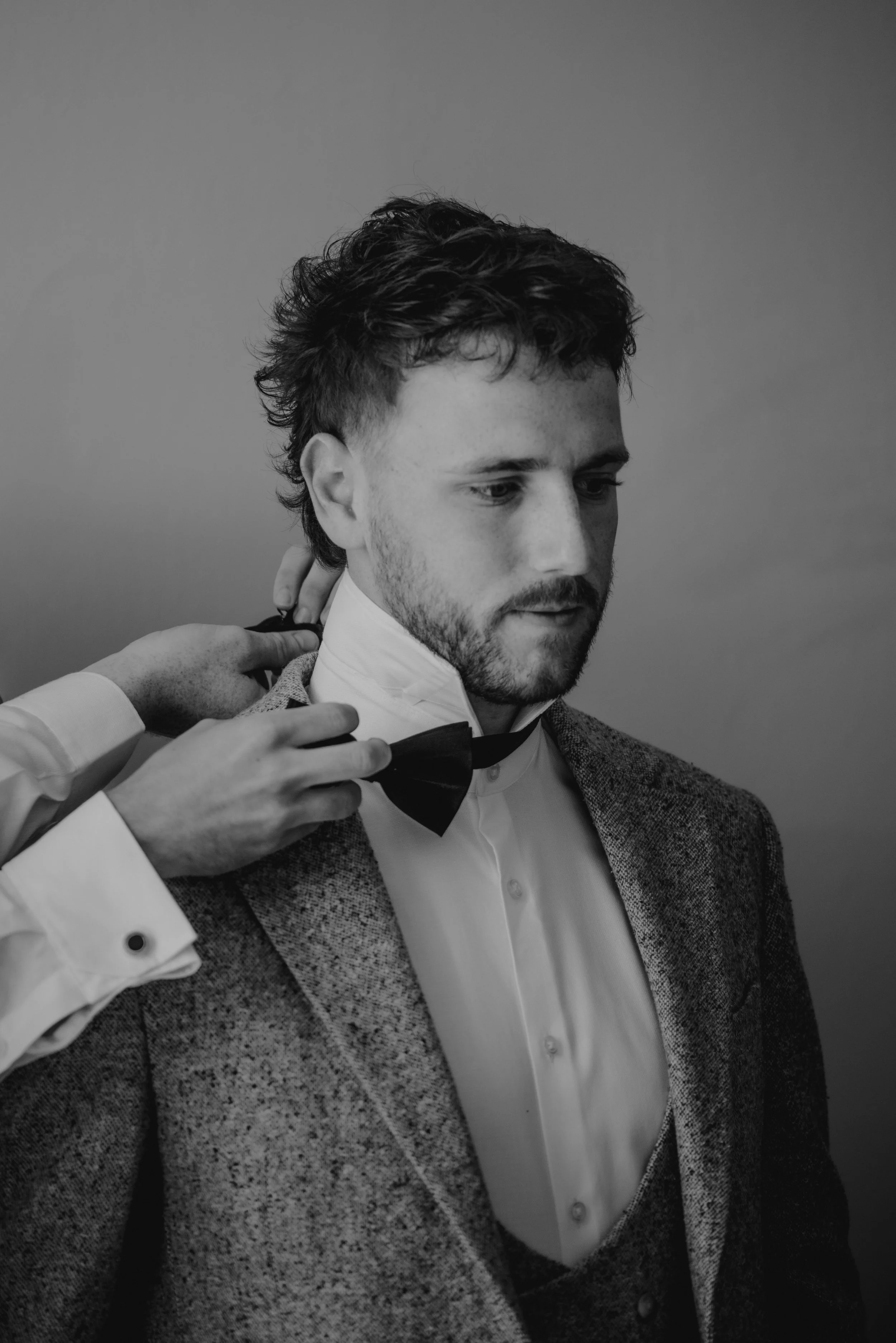 A man with styled hair and a beard is being helped to adjust his bow tie, wearing a tuxedo jacket and white shirt, against a plain background.