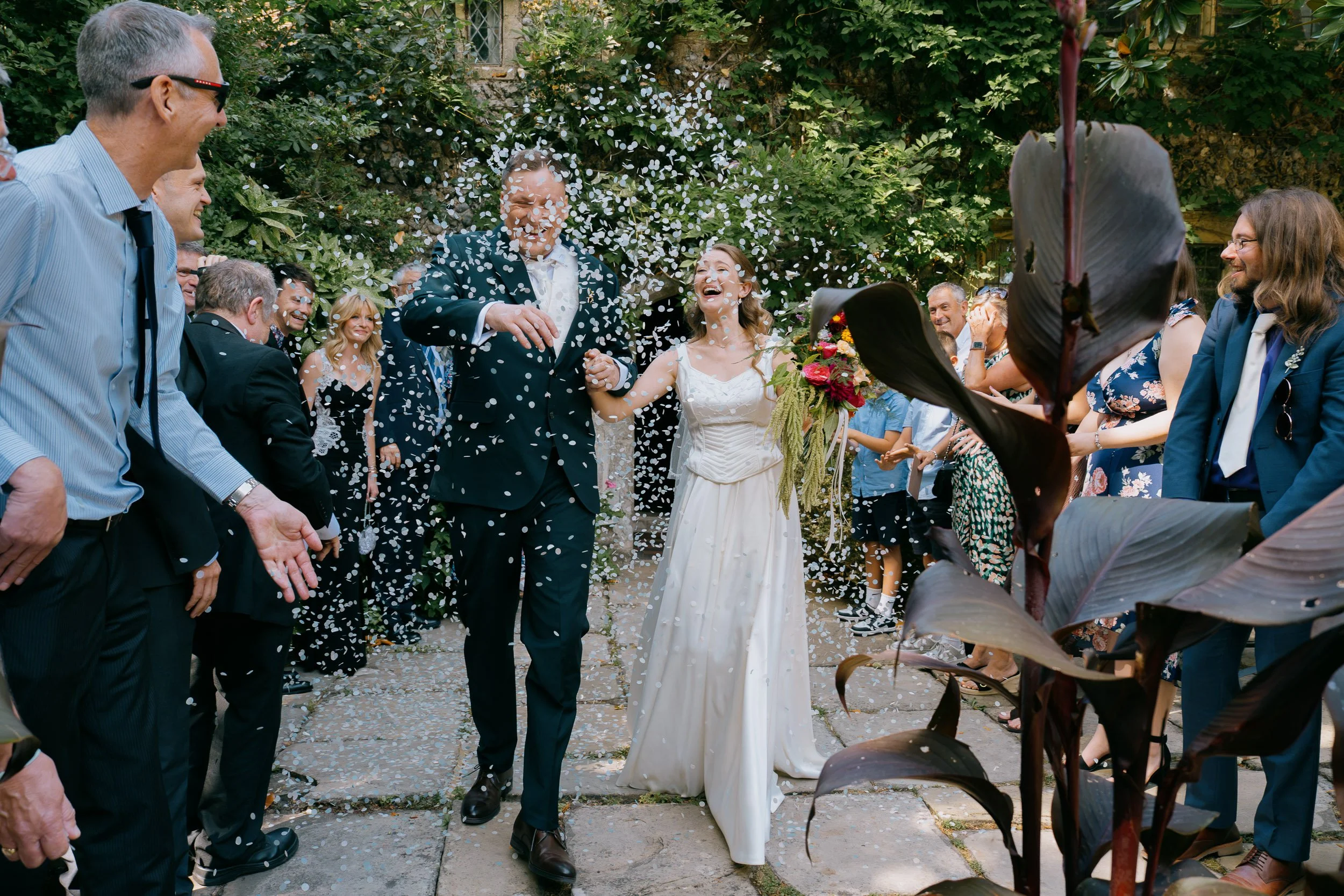 A bride and groom smiling and holding hands at their wedding reception, surrounded by friends and family, with confetti falling around them outdoors.