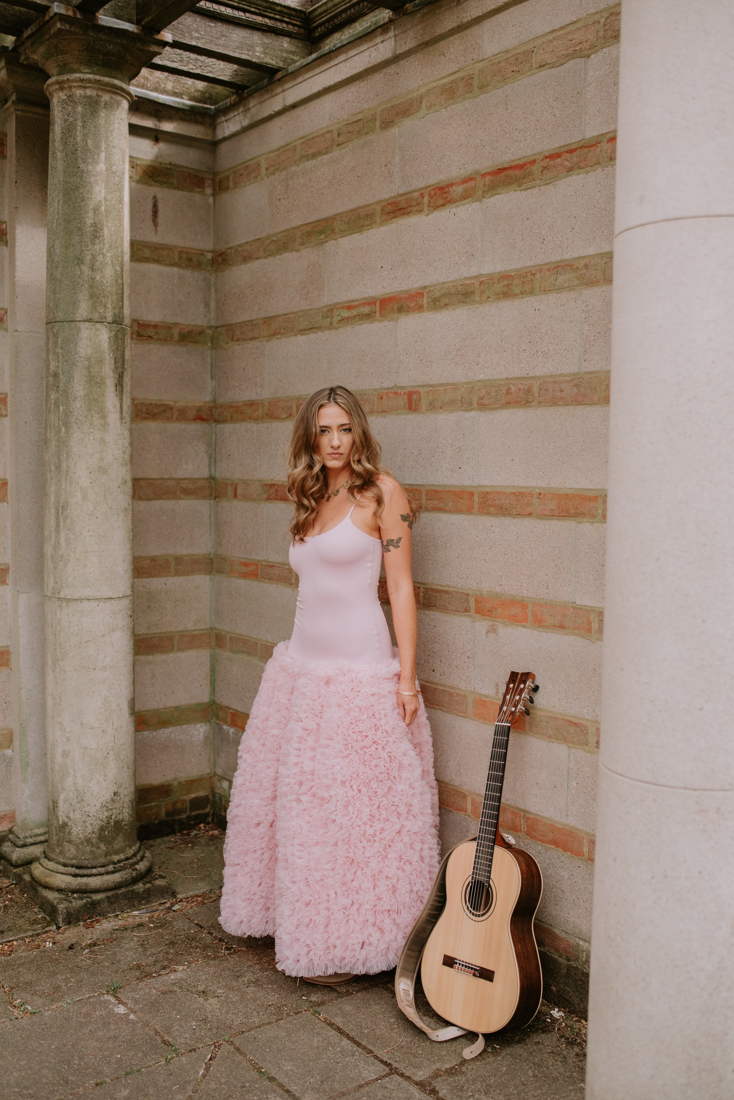 A young woman with wavy brown hair and tattoos on her arms stands next to an acoustic guitar leaning against a brick wall. She wears a pink dress with a fitted bodice and a fluffy textured skirt, in an outdoor setting with stone columns.