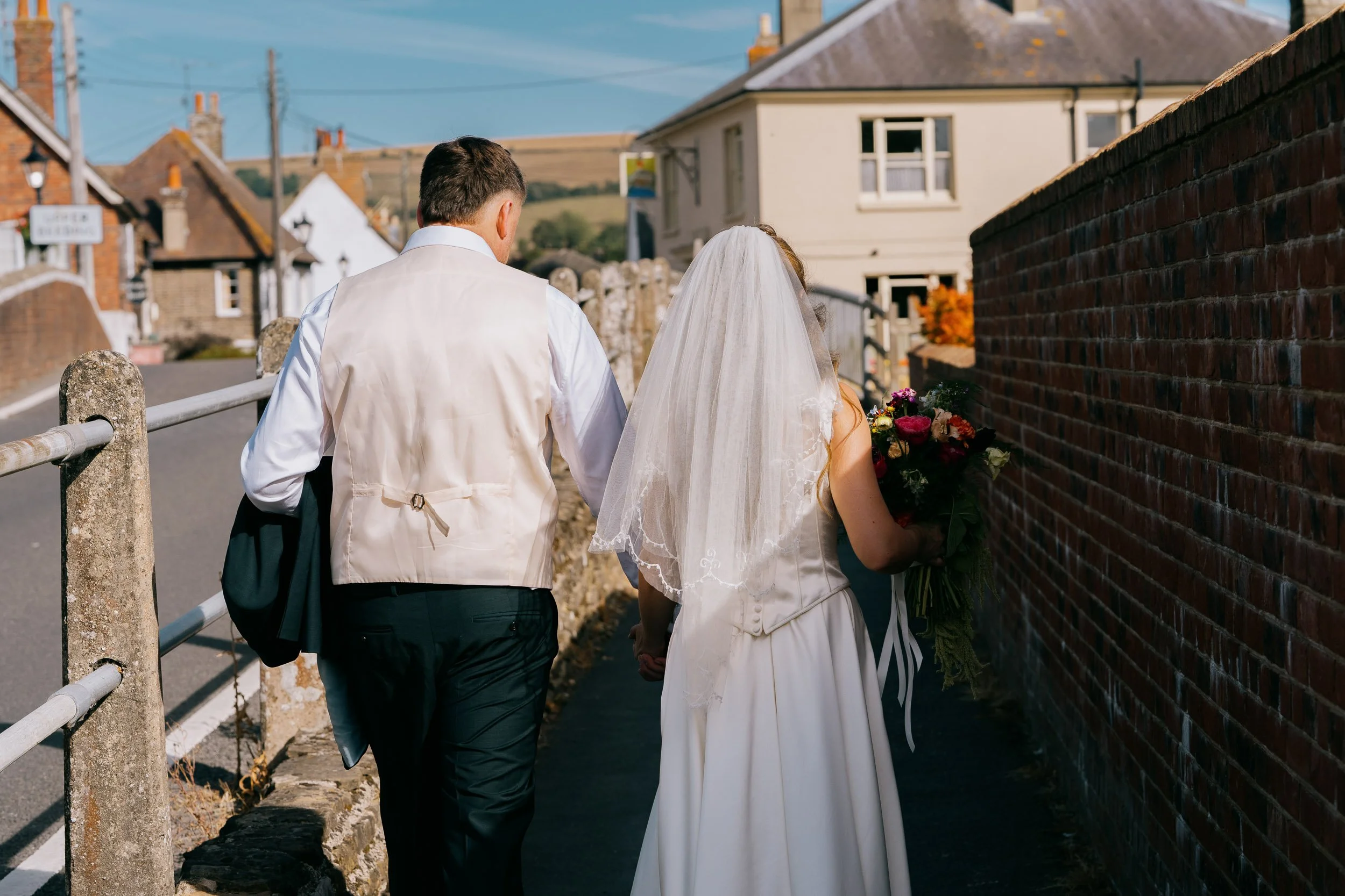 A bride and groom walking hand in hand in a rural neighborhood. The bride has a veil and holding a bouquet of flowers, and the groom is dressed in a white shirt with a beige vest. They are walking along a sidewalk with a brick wall on one side and ho