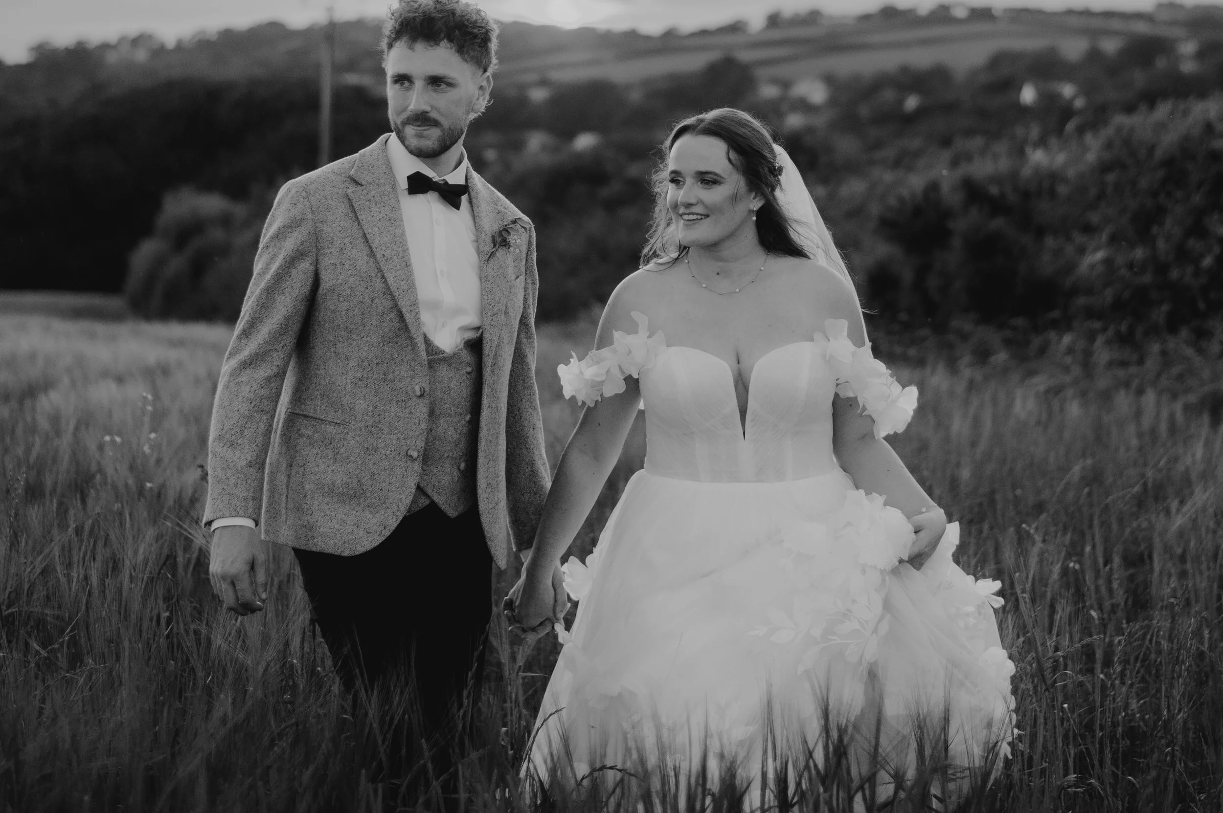 A black-and-white photo of a couple holding hands in a grassy field. The woman is in a wedding dress with off-the-shoulder floral details, and the man is in a suit with a bow tie. They are outdoors with a scenic cornwall countryside background.