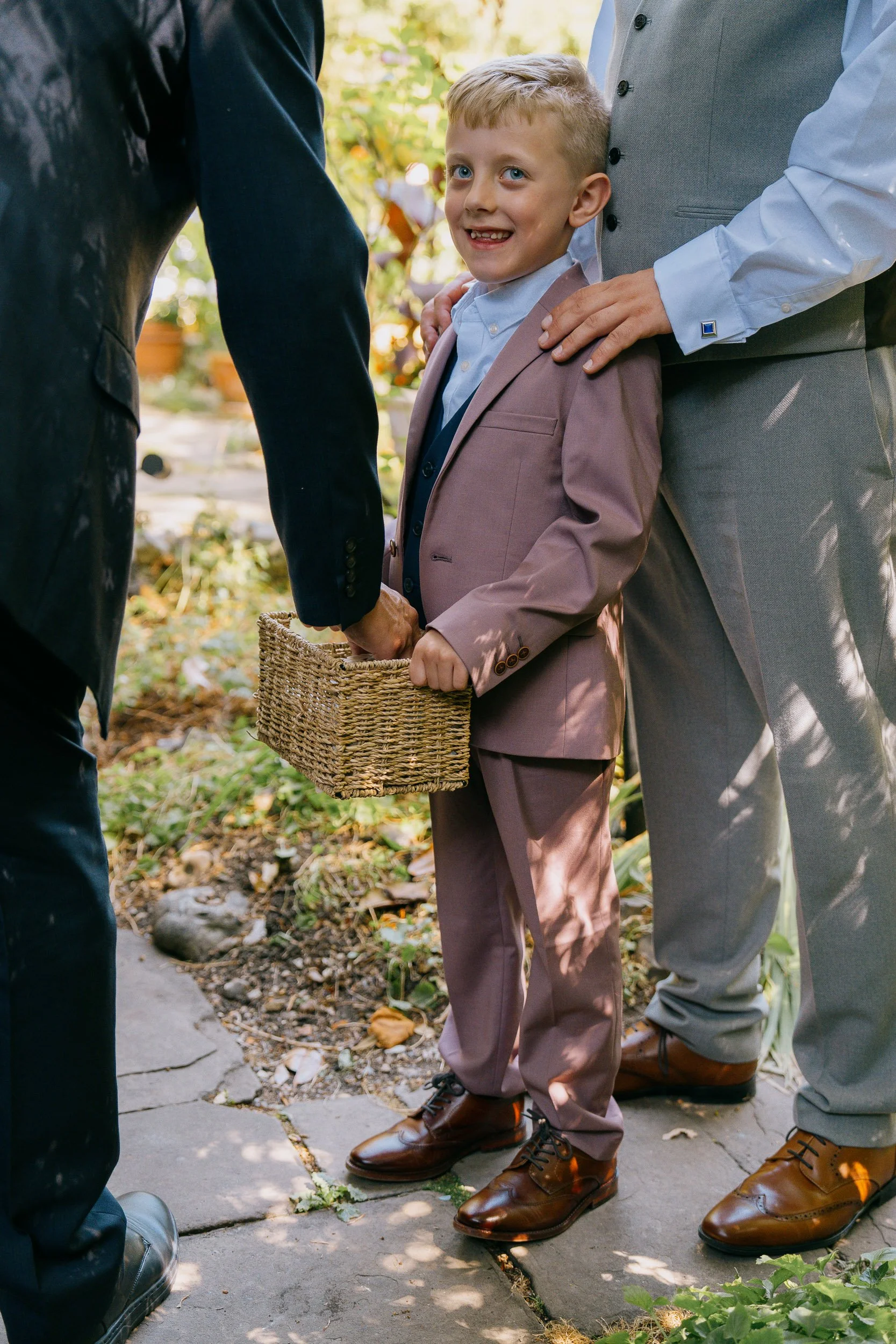 A young boy in a suit and tie holding a small woven basket, standing between two adults dressed in semi-formal attire, all outdoors on a sidewalk. The boy is smiling and looking at the camera.