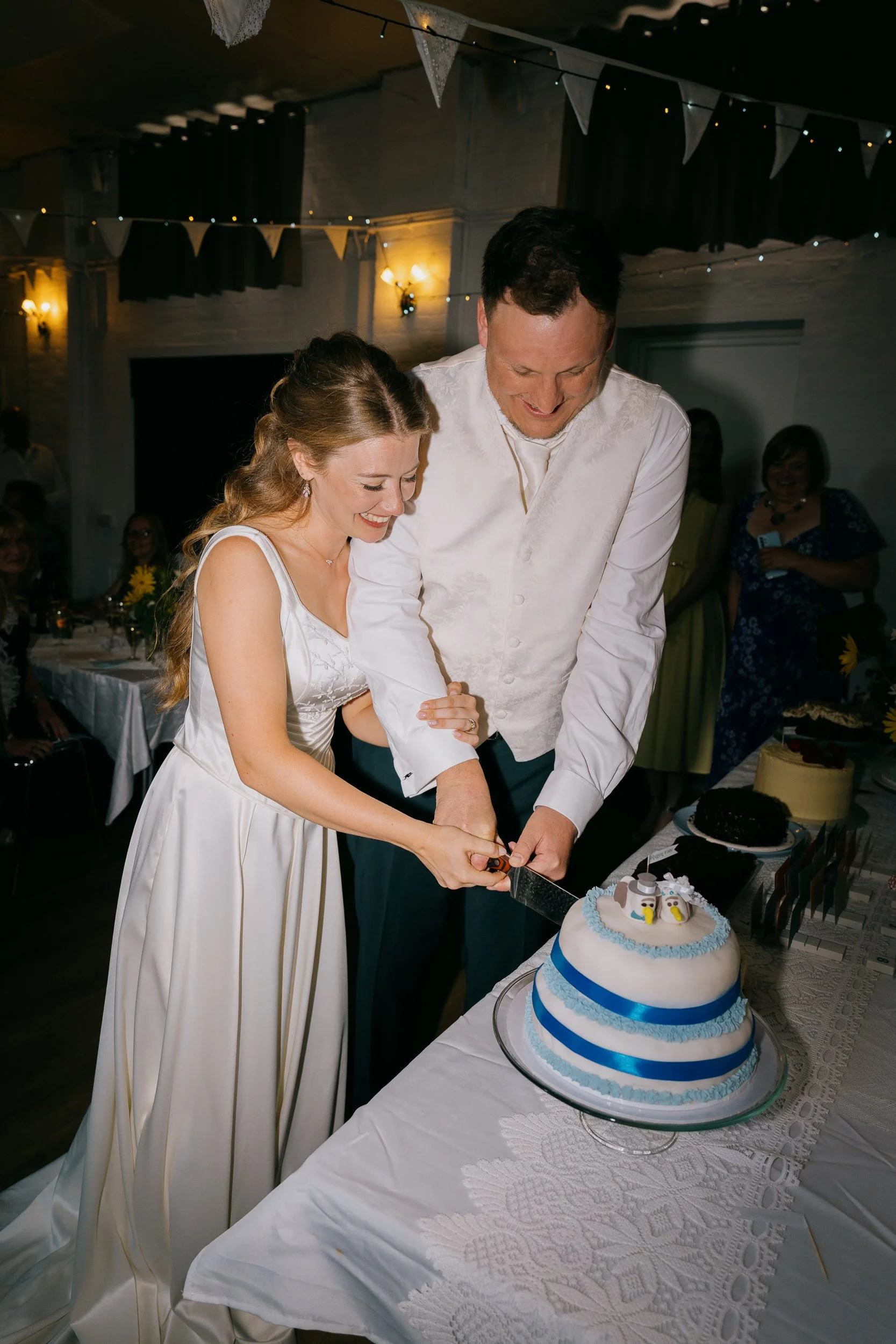 A bride and groom cutting a wedding cake at their celebration. The cake is decorated with blue ribbon bands and penguin toppers. Guests are seated in the background, and festive bunting and string lights decorate the room.