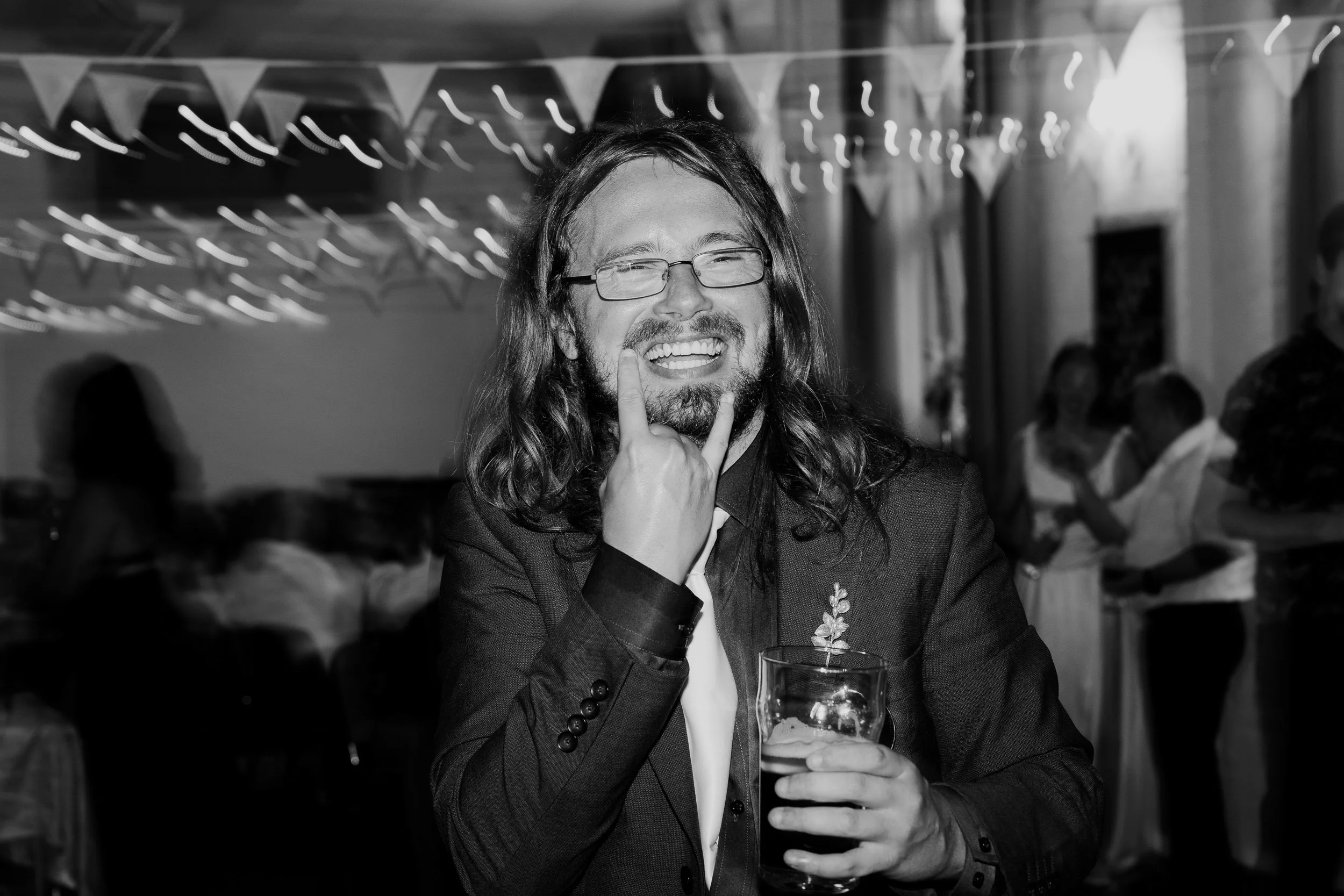 A man with long hair, glasses, and a beard smiling and making a peace sign with his fingers while holding a drink at a festive gathering.