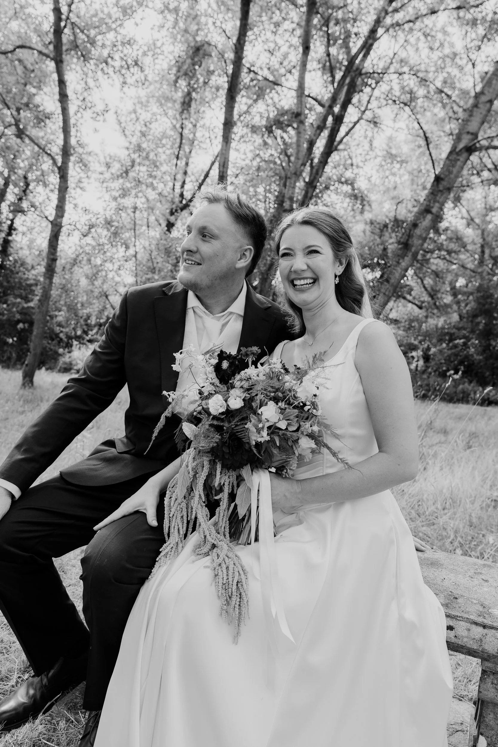 Black and white photo of a happy bride and groom sitting outdoors among trees, with the bride holding a bouquet of flowers, both smiling and looking to the left.