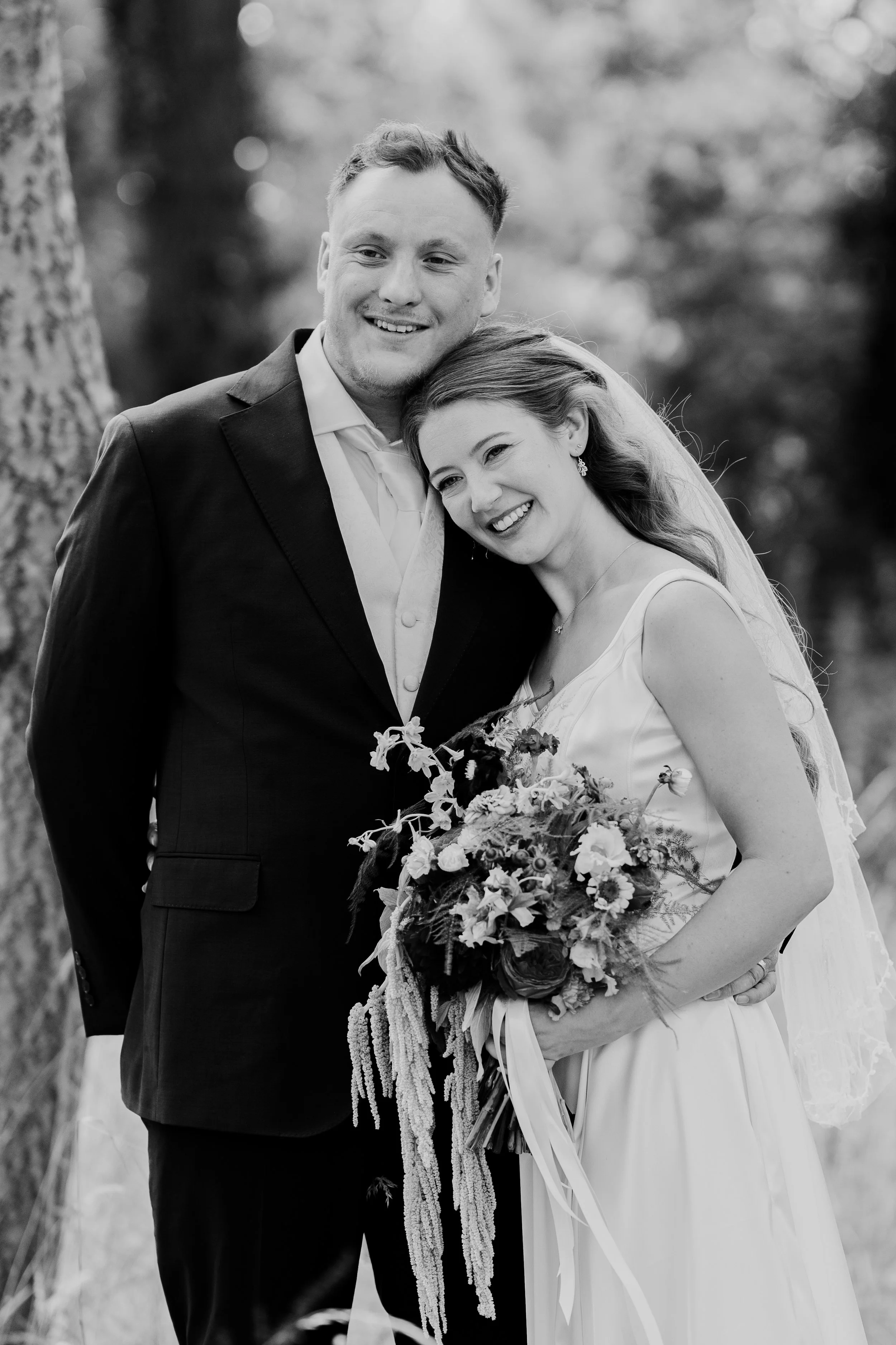 Black and white photo of a smiling bride and groom, outdoors, with trees in the background. The bride is holding a bouquet of flowers, leaning her head on the groom's shoulder, both dressed in wedding attire.