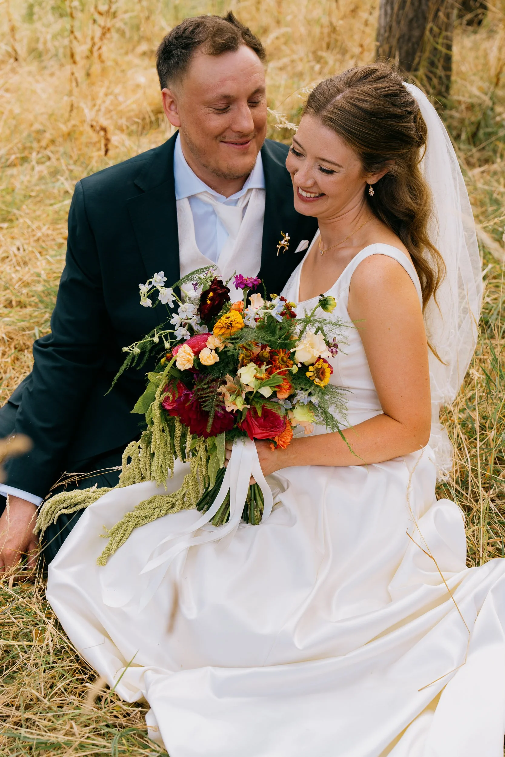 A bride and groom sitting in a field of dry grass, smiling and looking at each other, with the bride holding a colorful bouquet of flowers.
