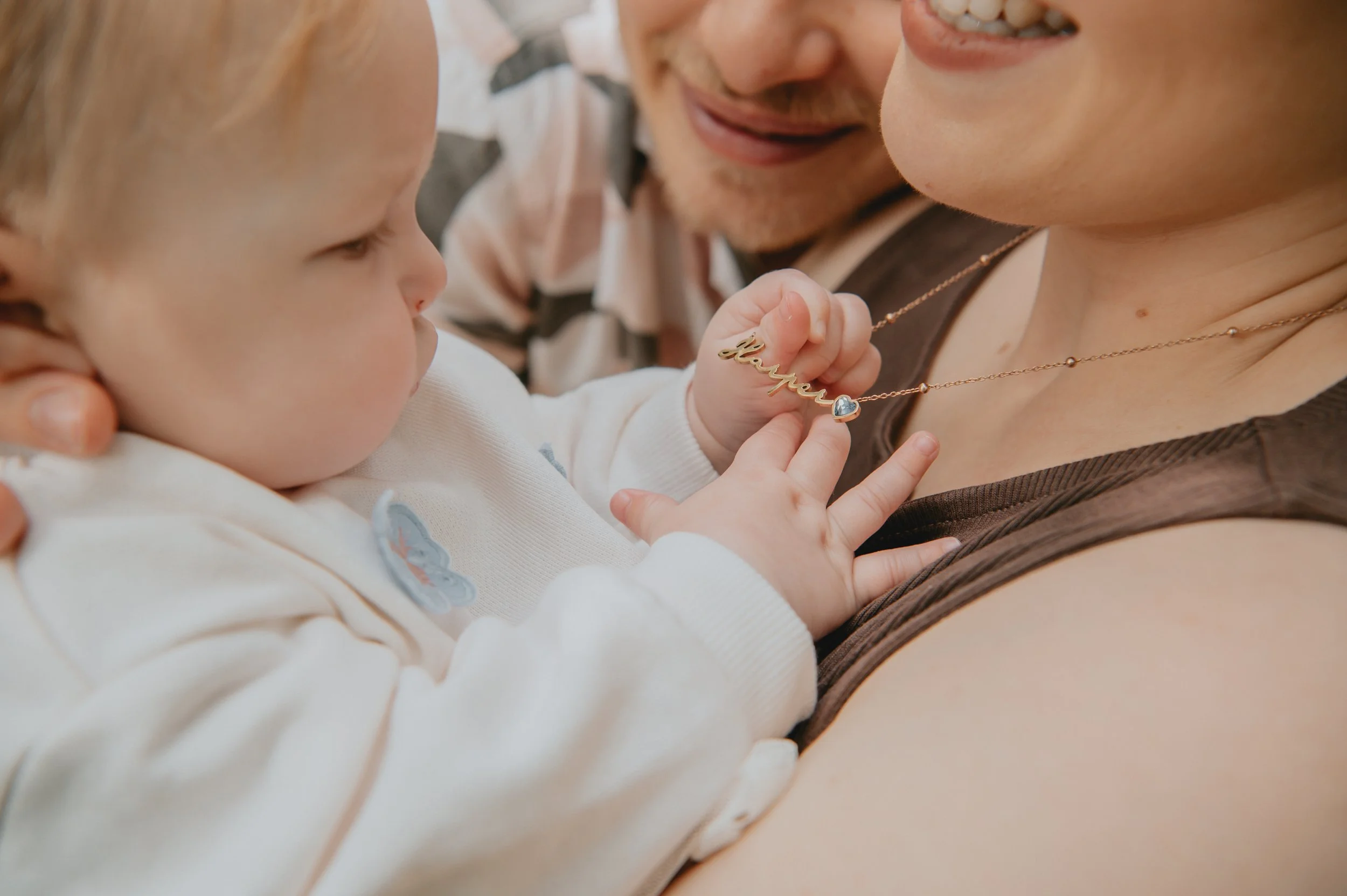 A young child and an adult sharing a tender moment. The child is holding a gold necklace with a name pendant and a heart-shaped charm, while the adult smiles warmly.