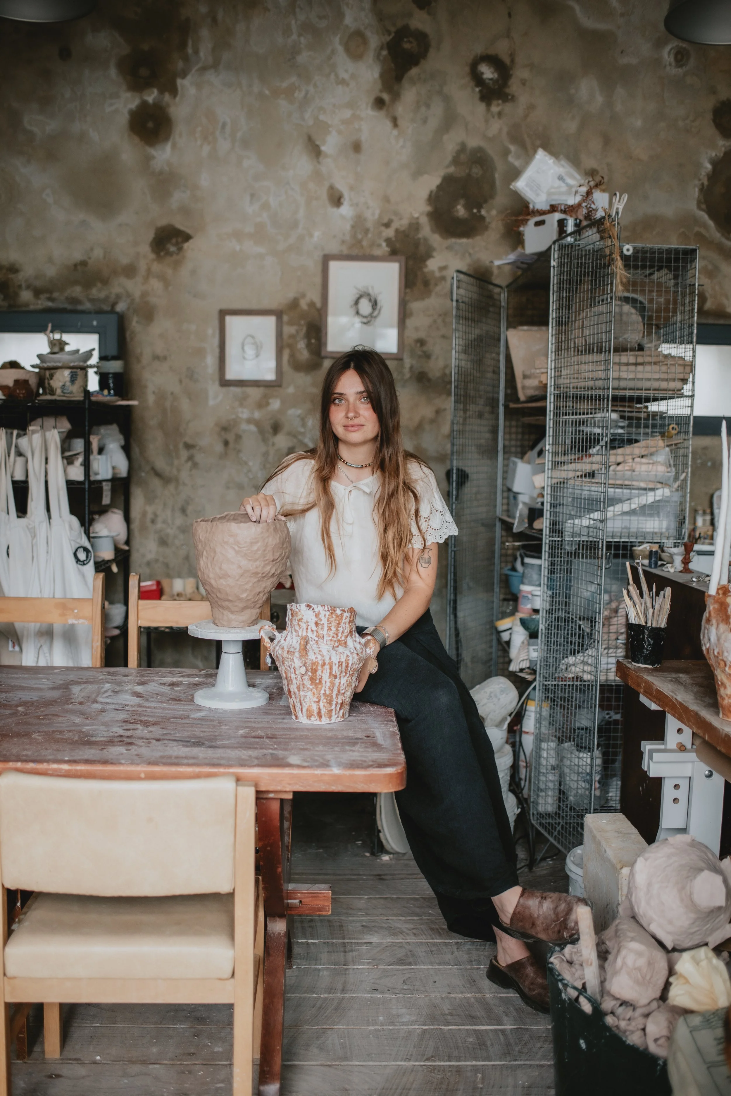 A young woman with long brown hair sitting in an art studio, with pottery on the table and shelves filled with art supplies in the background.