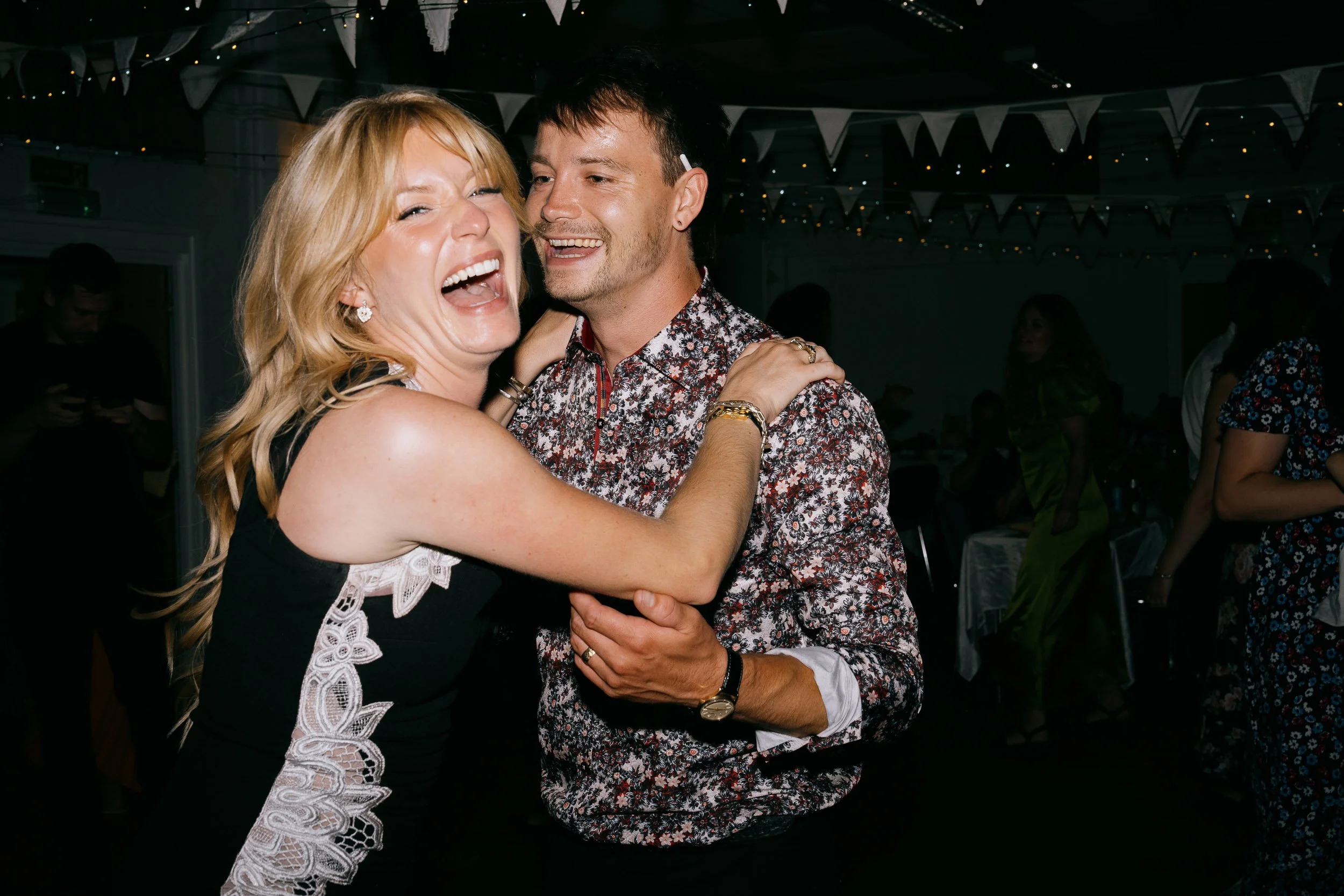 A woman with blonde hair and a man with dark hair are dancing and smiling at an indoor party with decorations and other guests in the background.