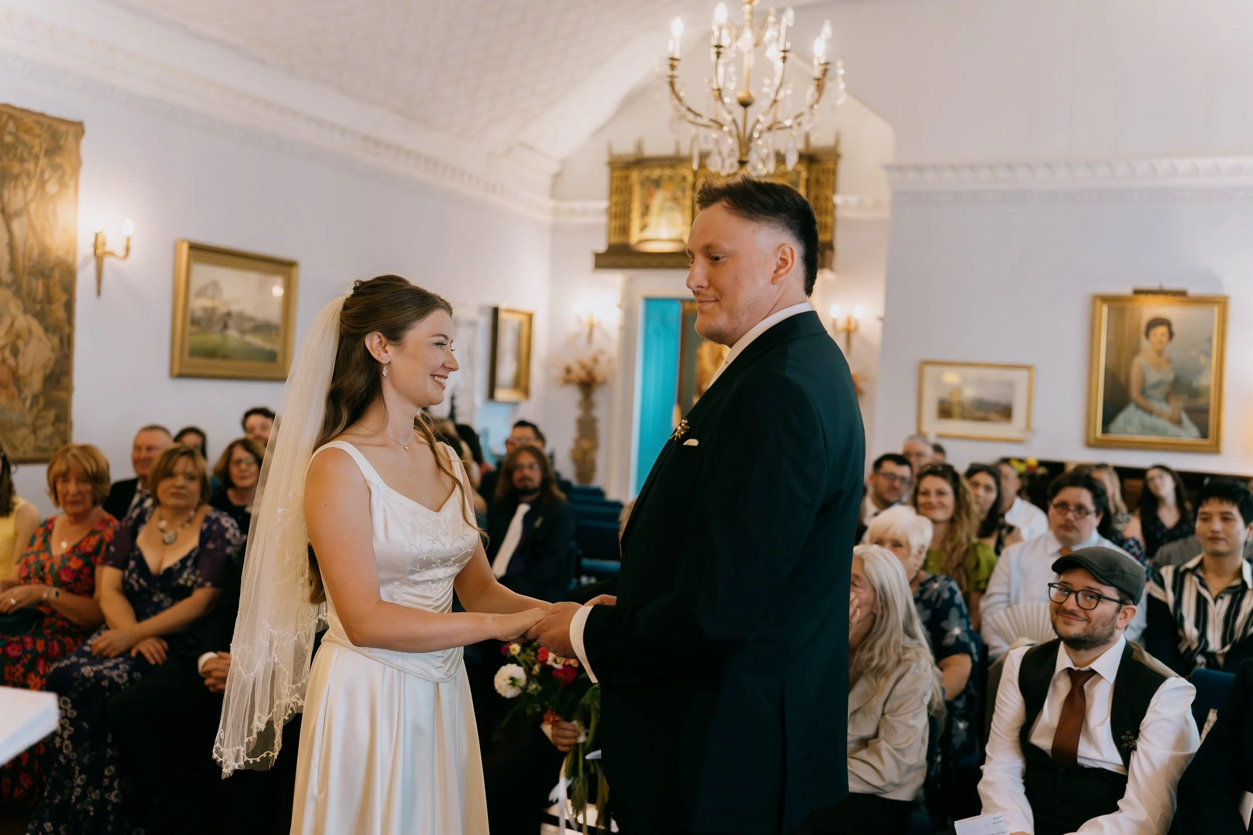 A bride and groom holding hands during their wedding ceremony in a decorated indoor venue with framed art on the walls and guests seated in the background.