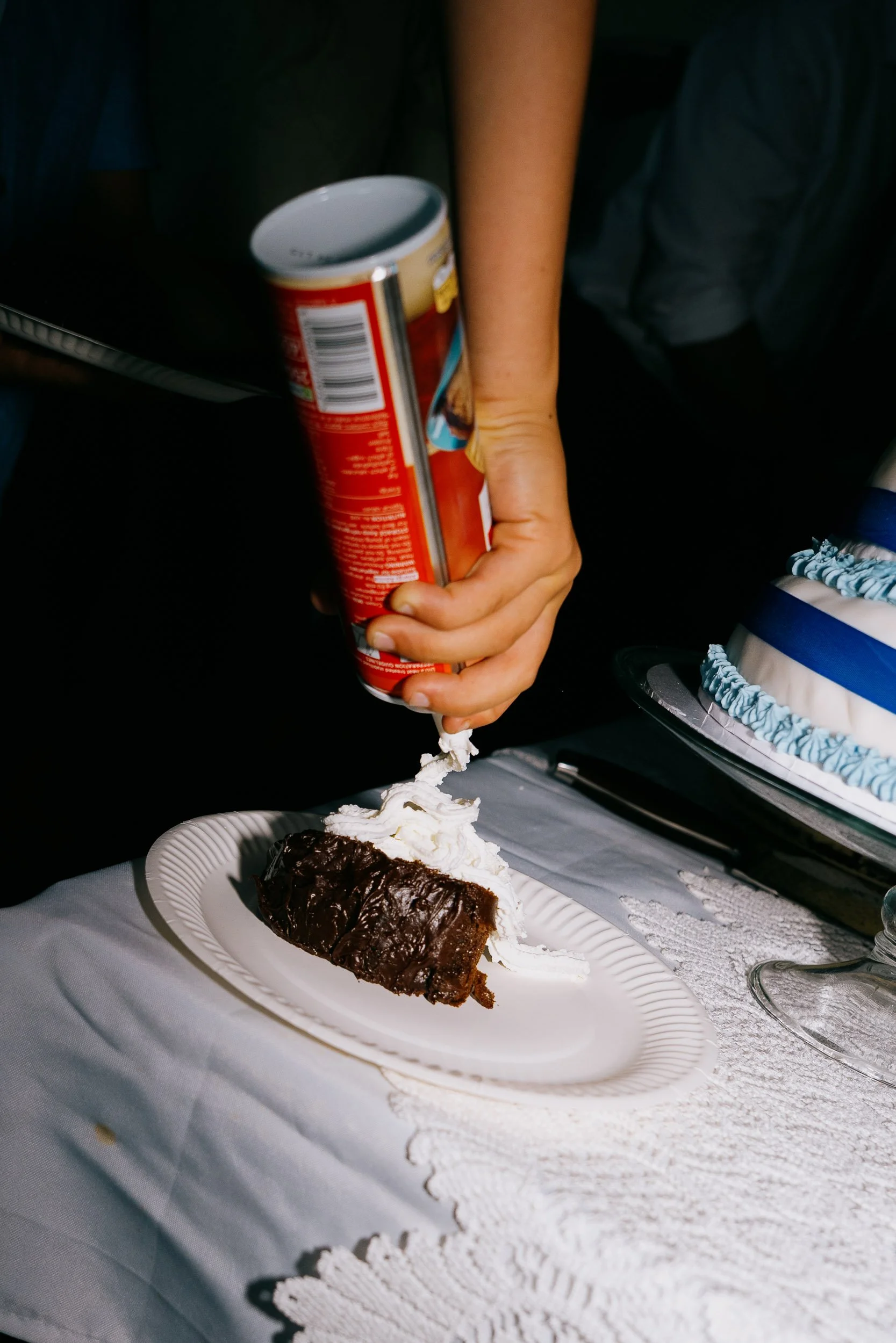 A slice of chocolate cake with whipped cream served on a white decorative plate on a white tablecloth, with a hand holding a can of whipped cream nearby.