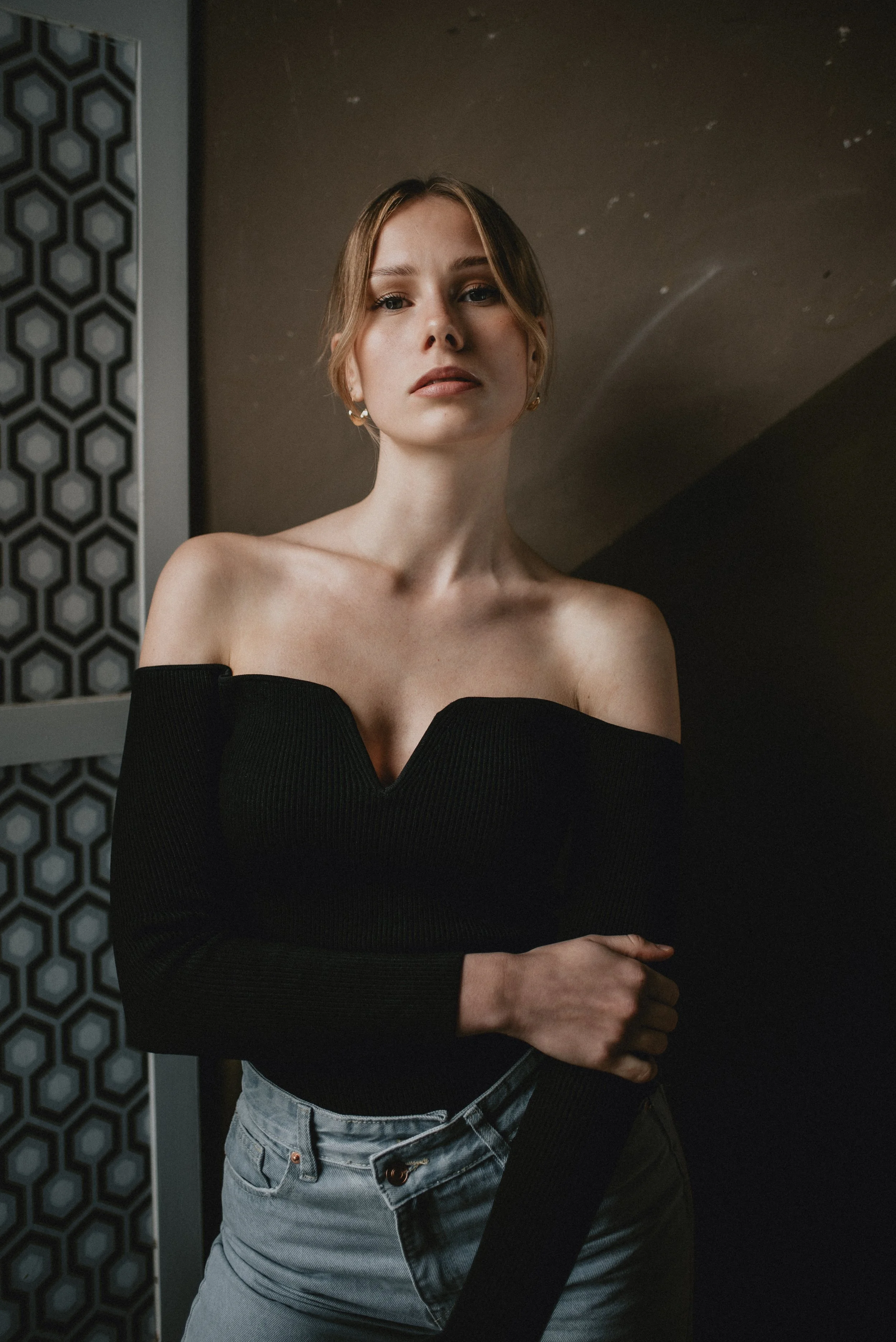 Young woman with fair skin and light brown hair in loose waves, wearing an off-the-shoulder black sweater and light blue jeans, standing indoors with a dark background and geometric patterned wall.
