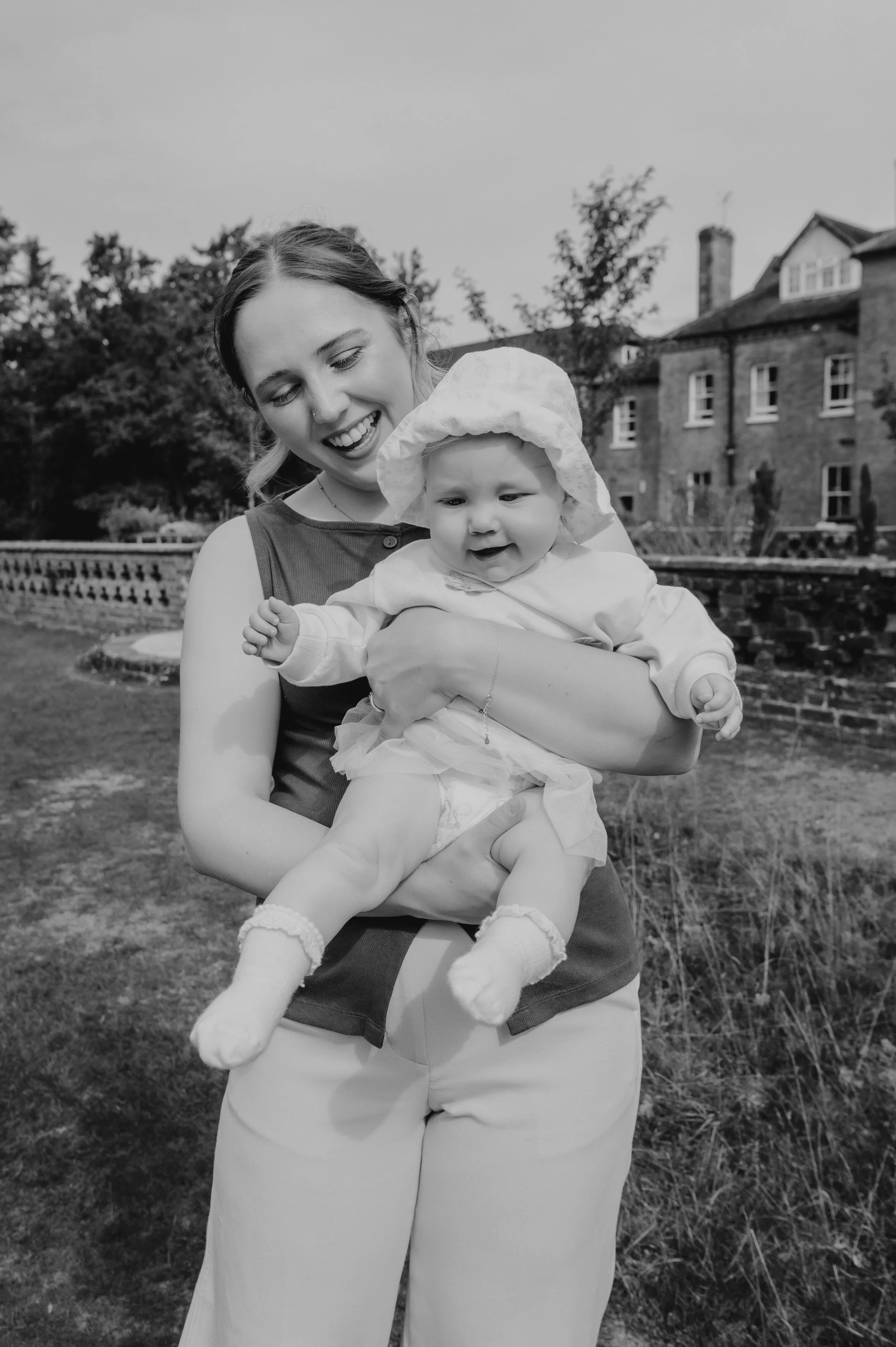 A woman holding a baby outdoors near a brick building and trees.