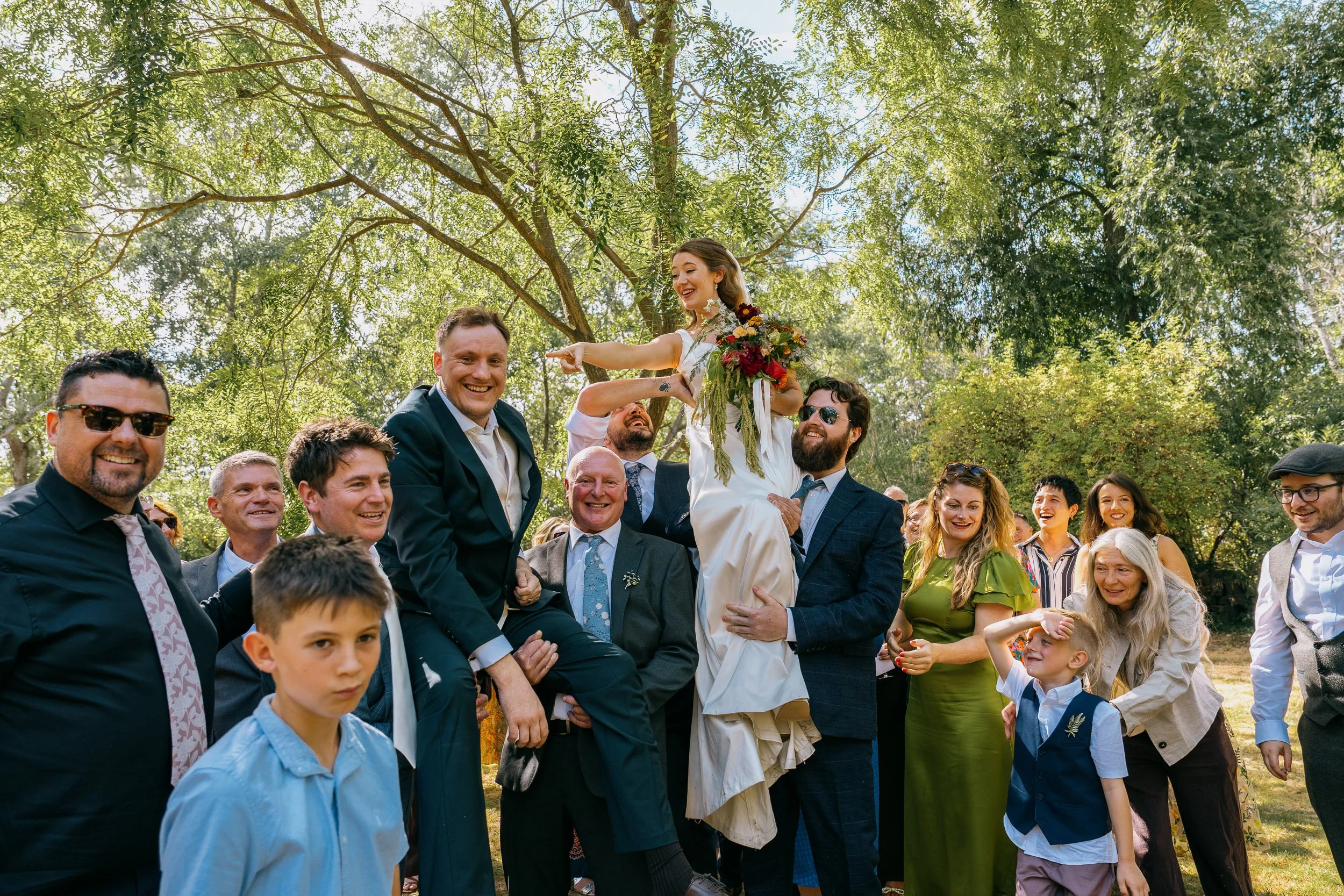 A wedding celebration outdoors with a bride and groom being lifted by friends and family, surrounded by lush trees and bright sunlight.