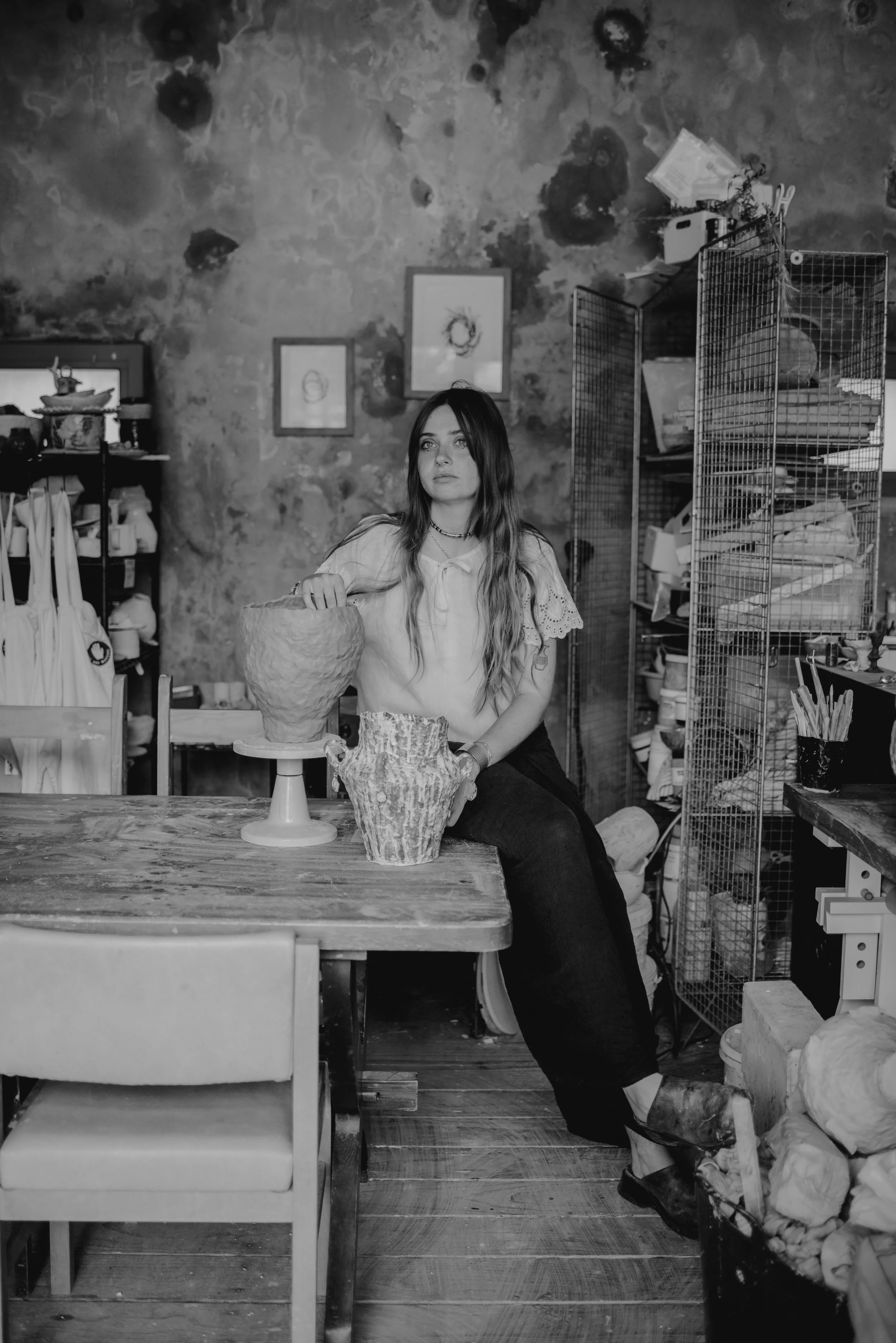 A woman with long hair sits at a wooden table in an art studio, surrounded by pottery and art supplies, with a textured wall and framed sketches behind her.