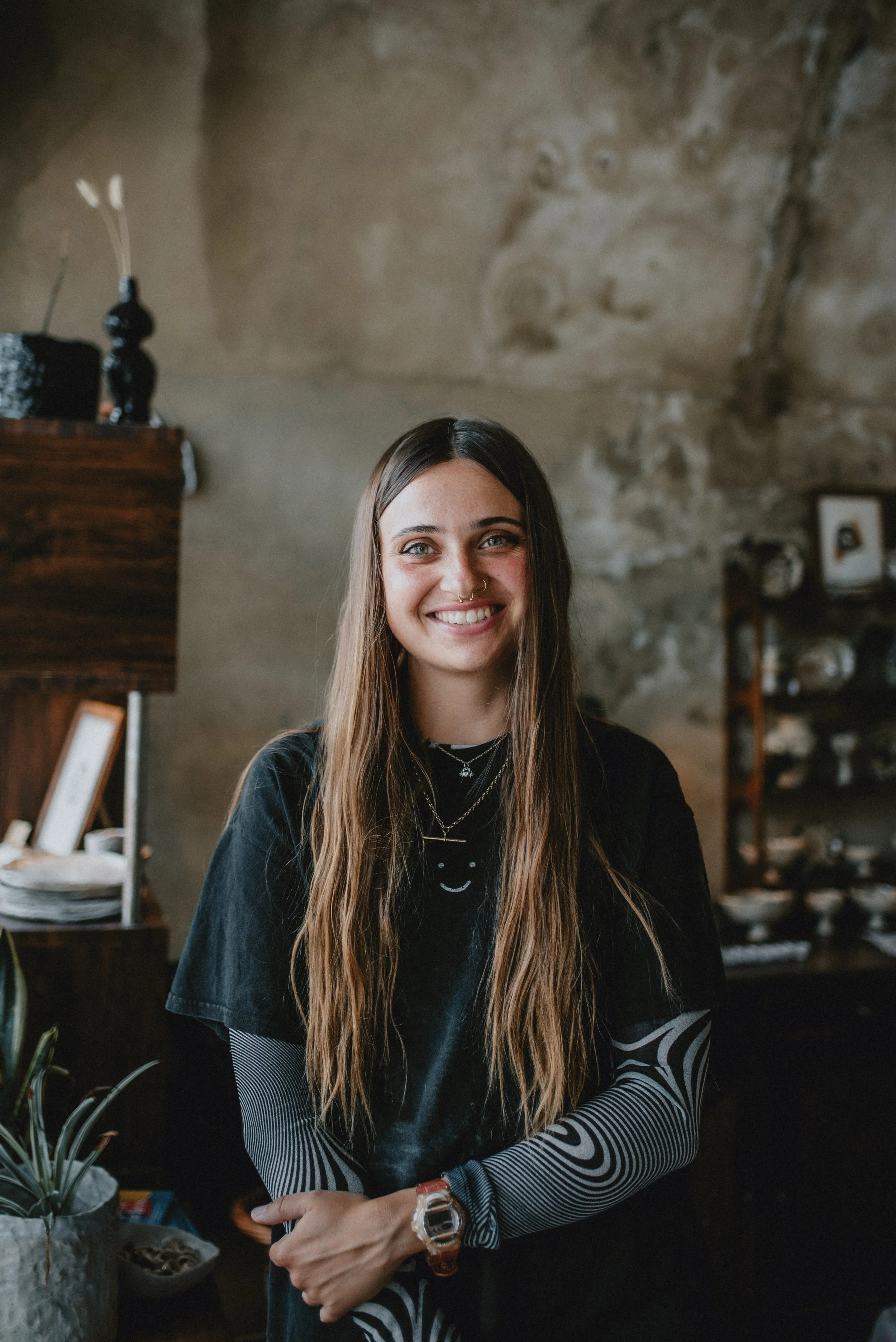 A young woman with long brown hair, smiling, wearing a black T-shirt with a white smiley face, layered necklaces, and a watch. She is standing indoors in front of a rustic stone wall, with shelves and decorations behind her.