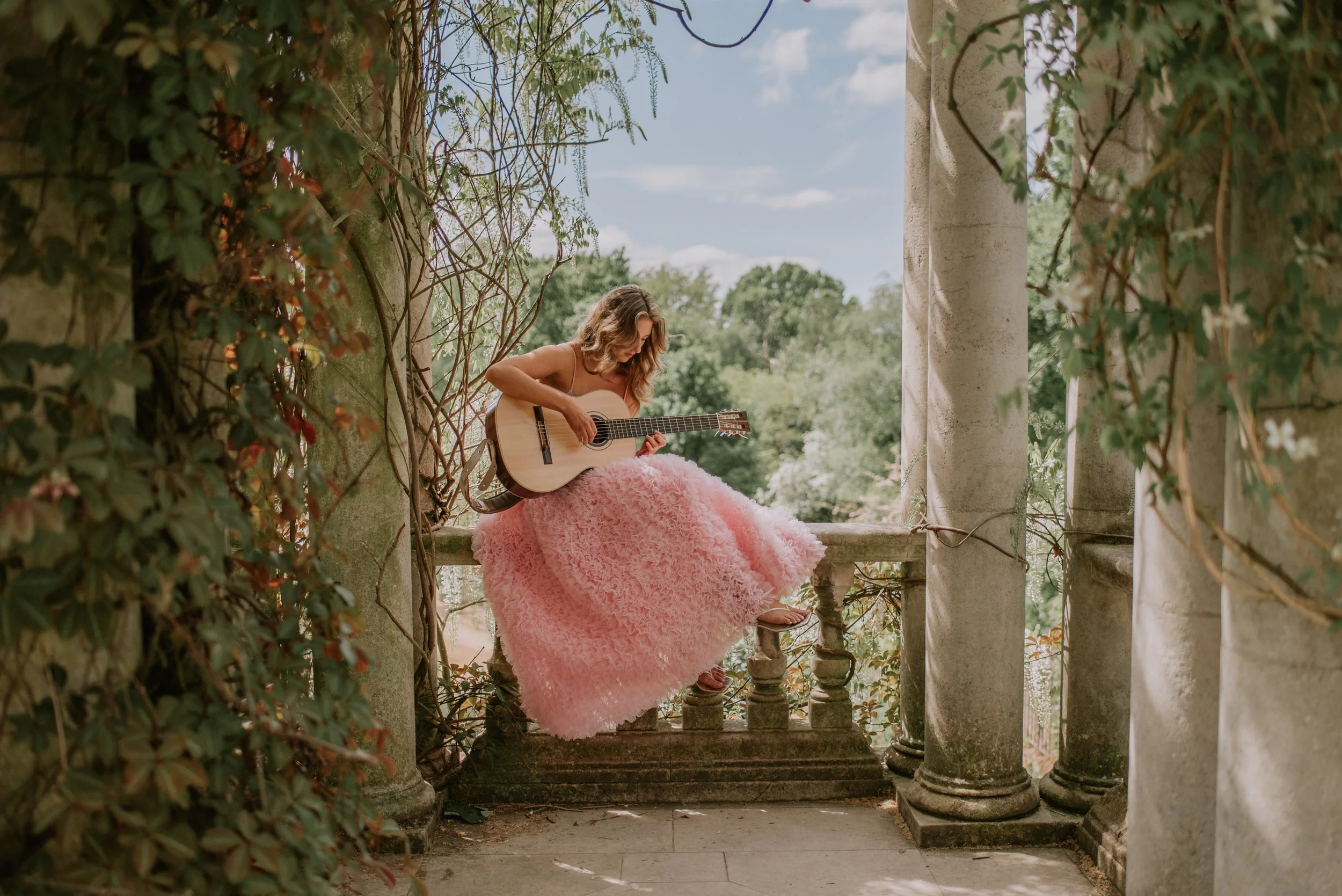 A woman in a pink, frilly dress sits on a stone ledge of an old stone and columned gazebo, playing an acoustic guitar. She is outdoors with lush green trees and a blue sky in the background.