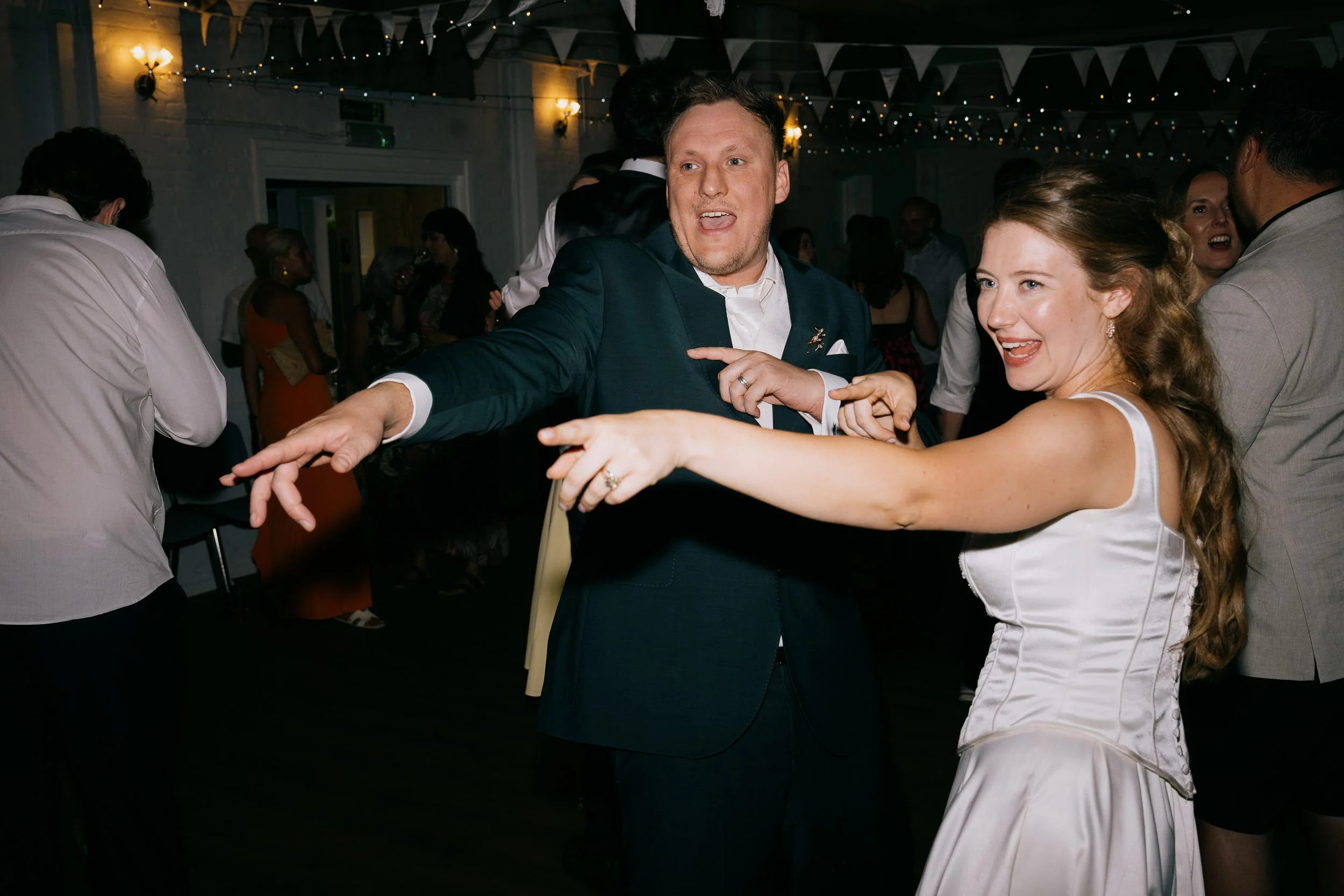 A man in a navy blue suit and a woman in a white dress are dancing and smiling at a party or celebration event. The background features other people and decorative string lights and bunting.