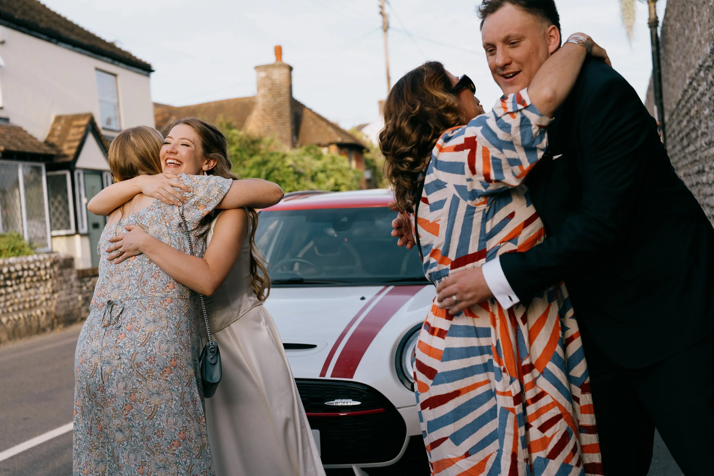 Four people hugging on a street, with two women on the left and a man and woman on the right. They are near a white car, and houses are visible in the background.