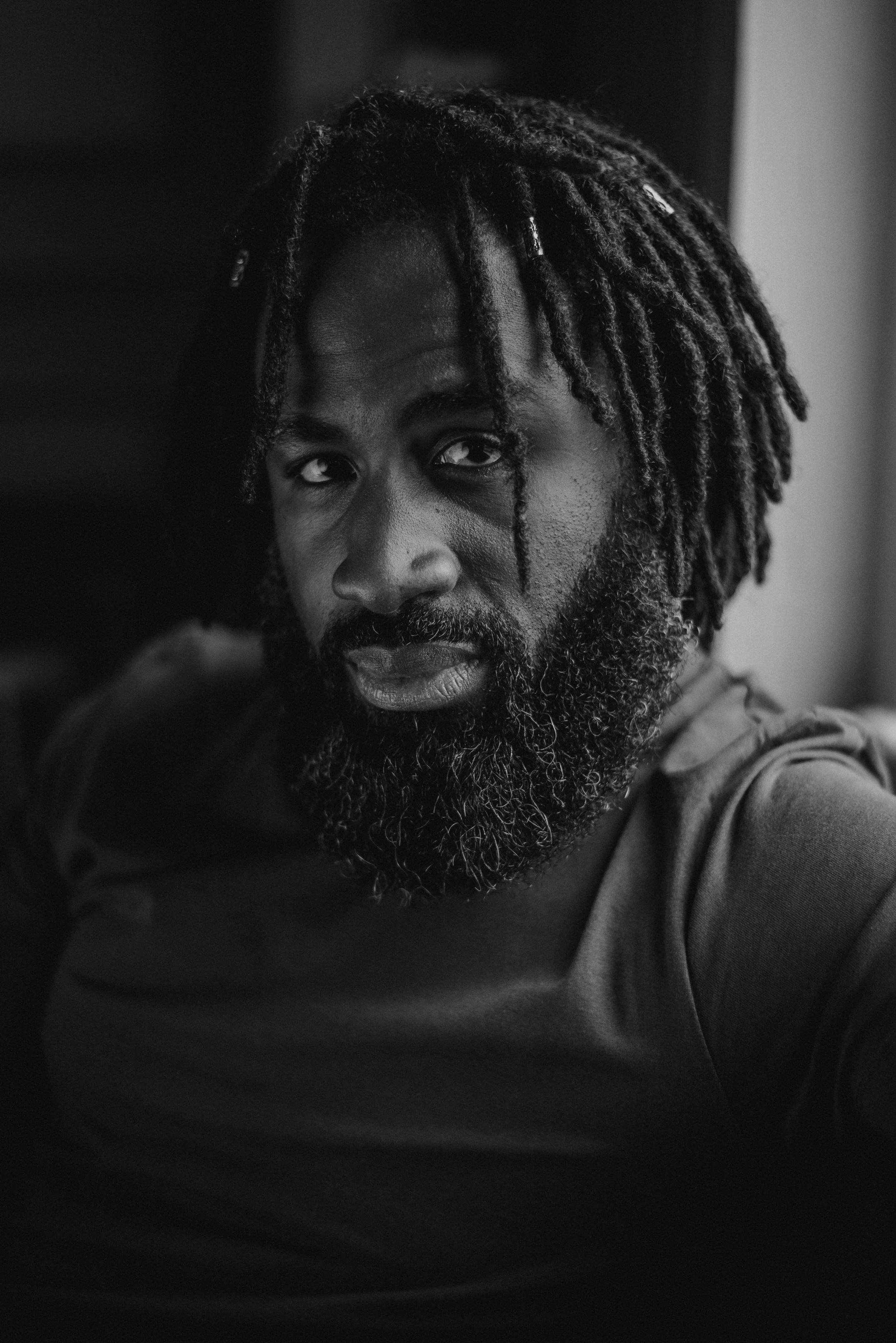 Black and white close-up portrait of a man with dreadlocks and a full beard, looking at the camera with a serious expression.