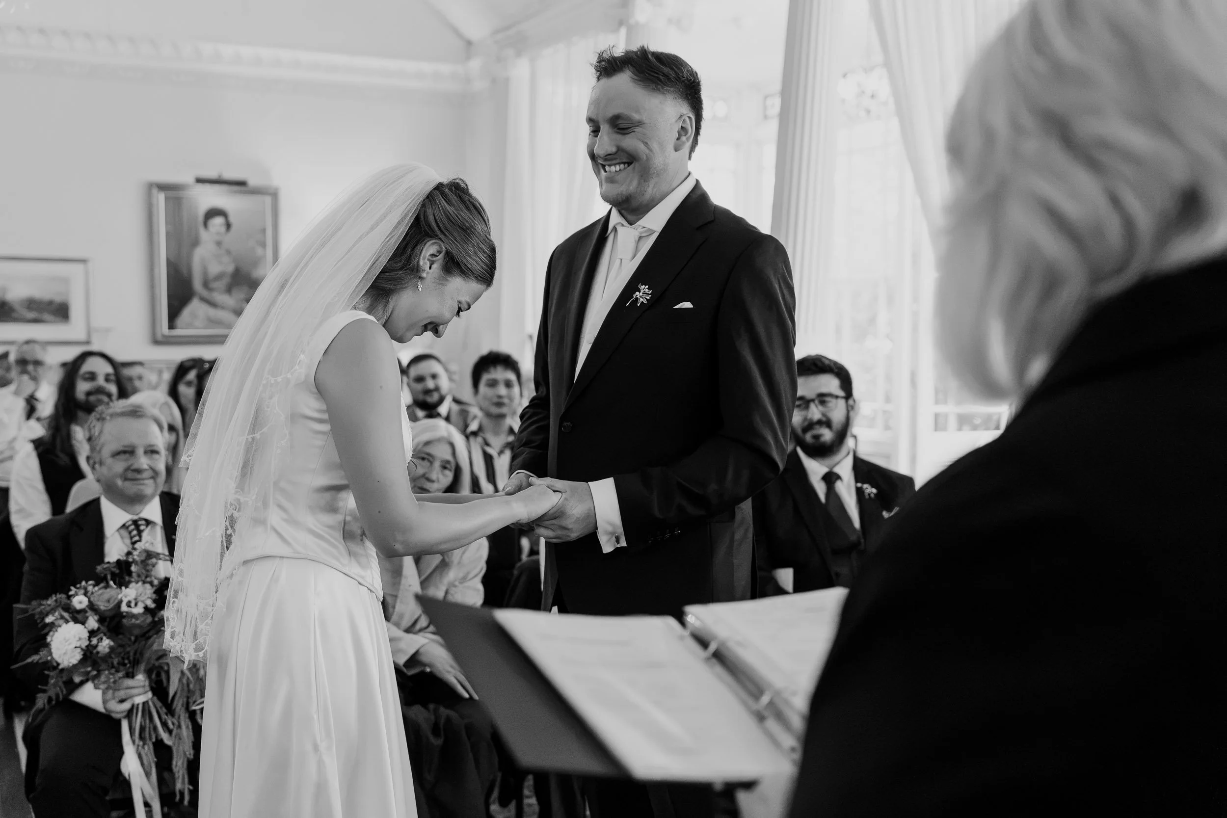 A black and white photo of a wedding ceremony showing a bride and groom holding hands and smiling. The bride wears a white dress and veil, the groom wears a dark suit. Guests are seated, watching happily. The setting appears to be indoor with large w