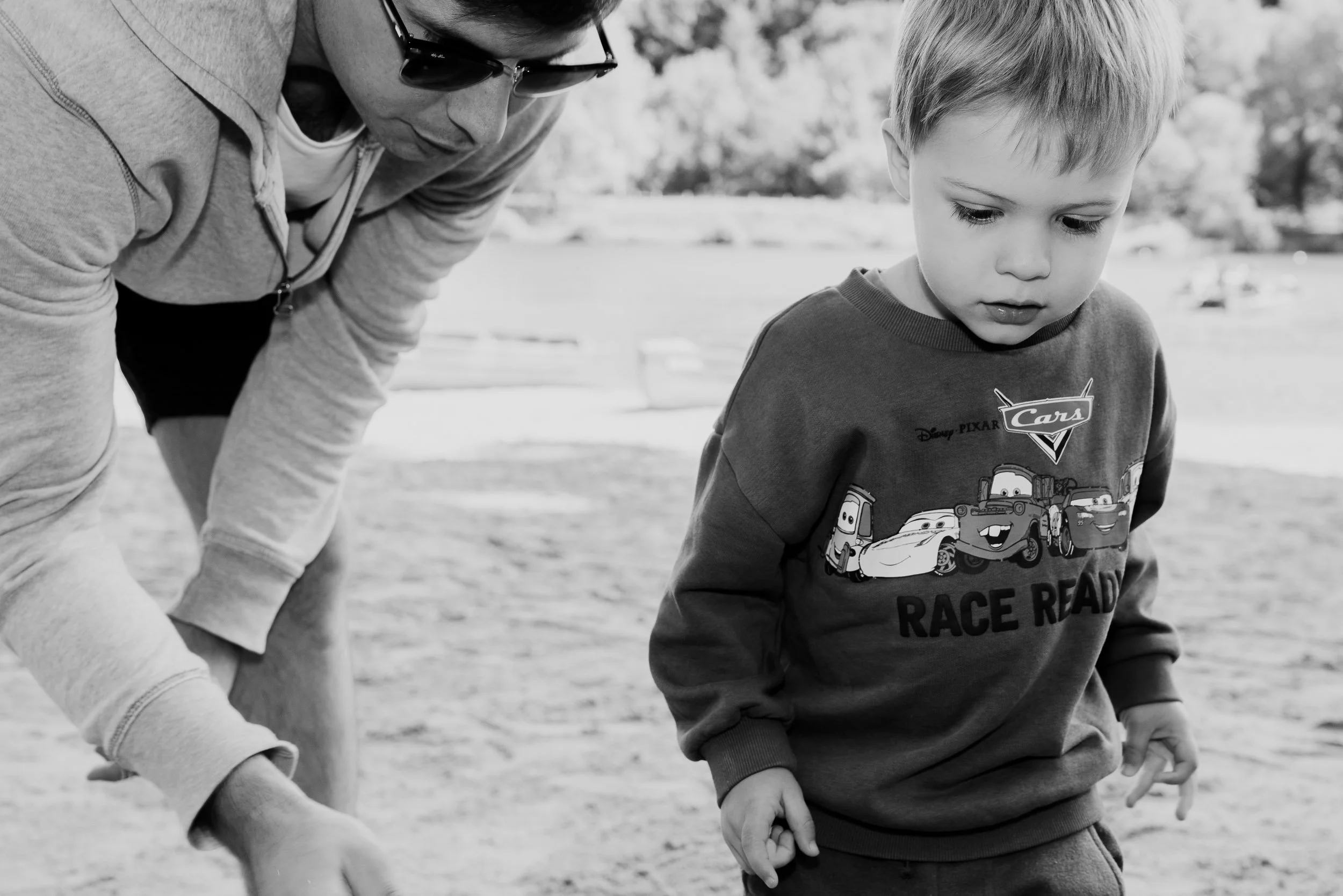 A man and a young boy are looking down at something outside on a cloudy day. The boy is wearing a "Cars" themed sweatshirt, and the man is wearing sunglasses and a hoodie.