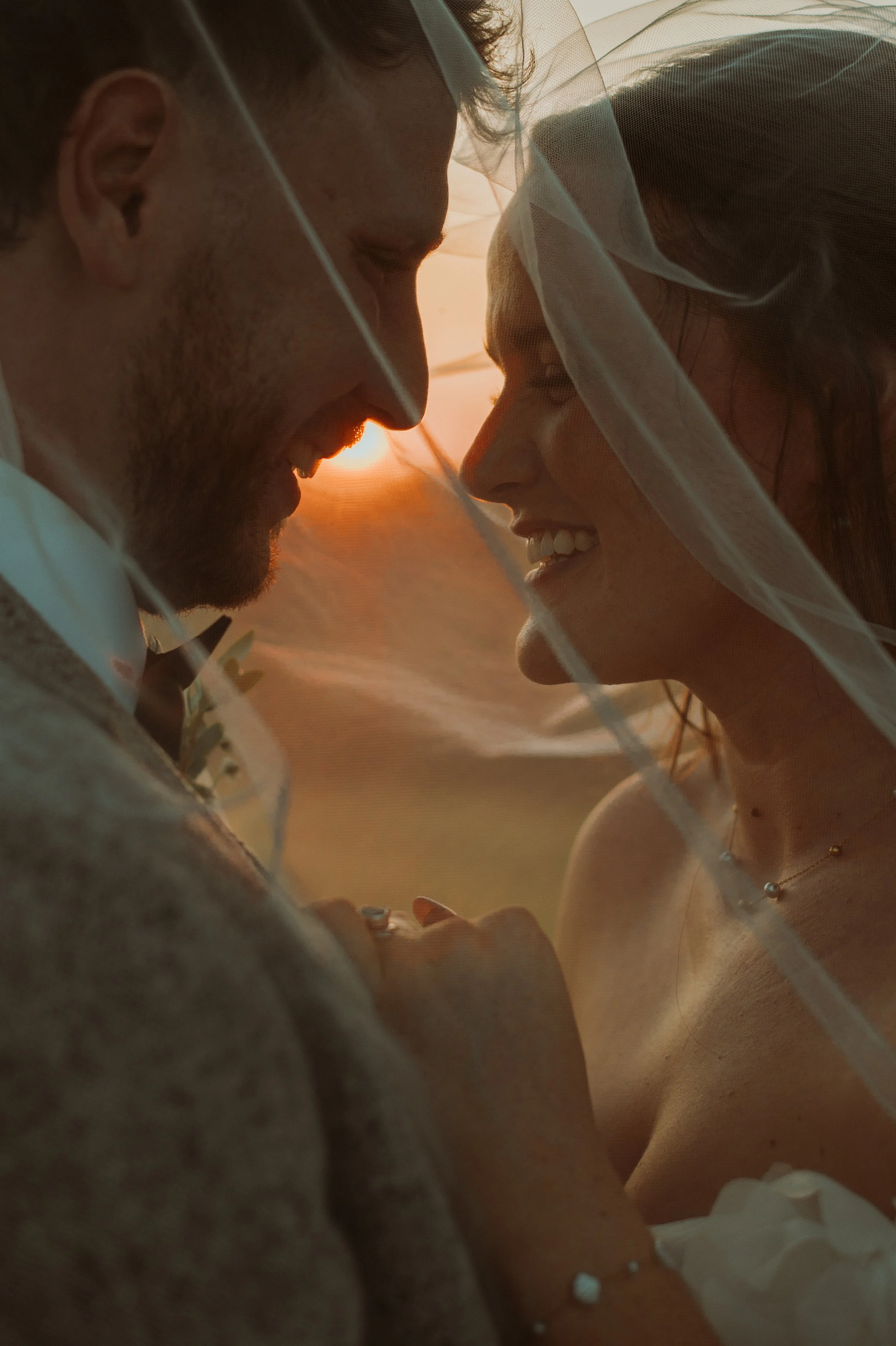 A man and woman smiling with foreheads touching, partially covered by a veil, during sunset.